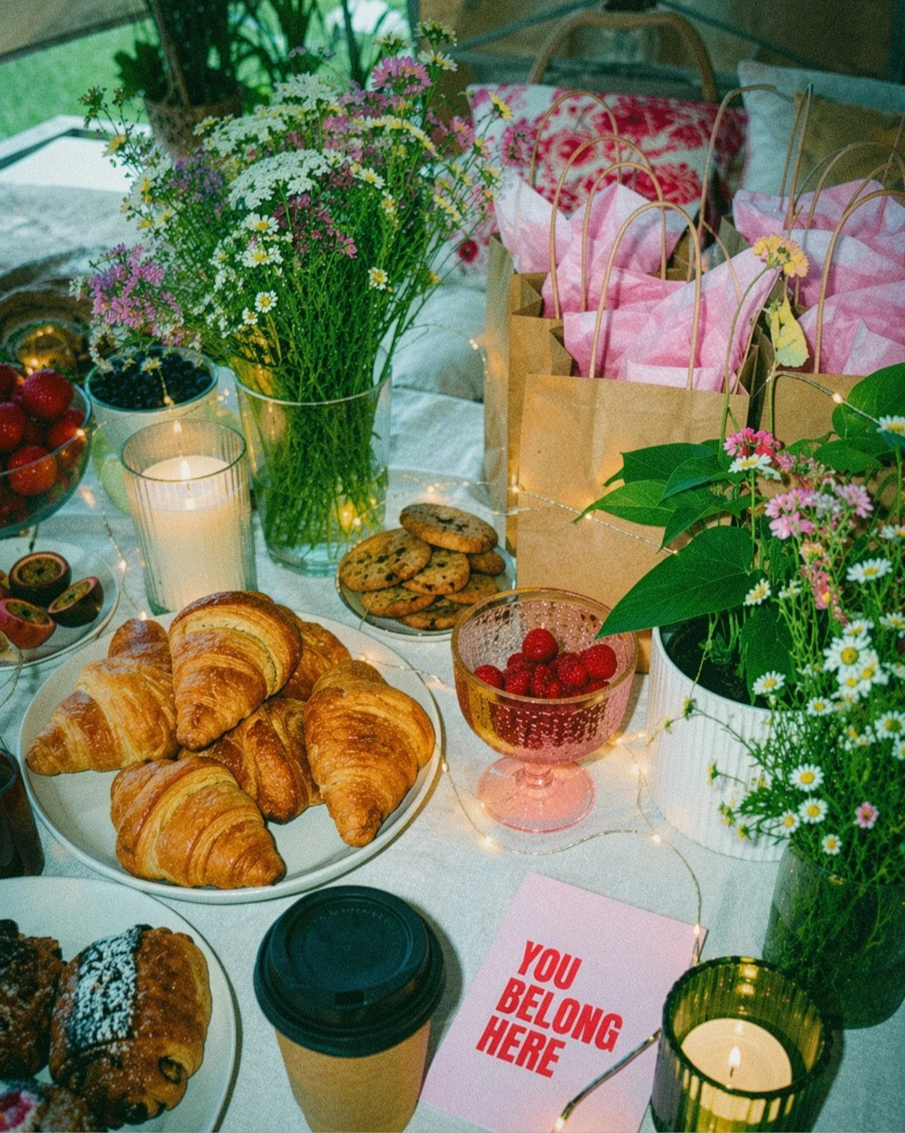 A table set with breakfast items including croissants, cookies, and pastries, surrounded by pink, yellow, and white flowers, candles, and cups of coffee, with gift bags and a sign that reads 'You Belong Here'.