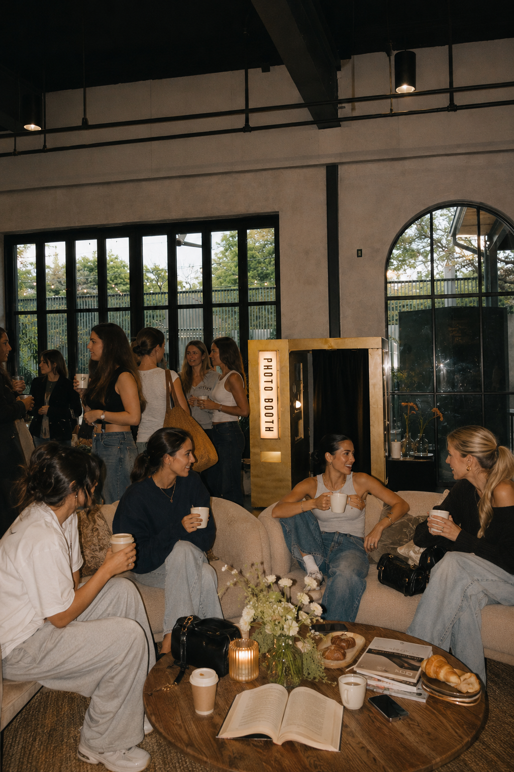 Group of women socializing and enjoying drinks at an indoor gathering, with a photo booth in the background and a table with flowers, books, and pastries in the foreground.