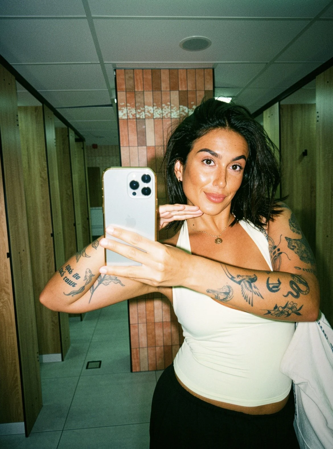 A woman with dark, wavy hair and tattoos on her arms is taking a mirror selfie in a bathroom. She is smiling and holding her phone with her left hand while her right hand is resting under her chin. She is wearing a sleeveless, light-colored top and has a necklace.