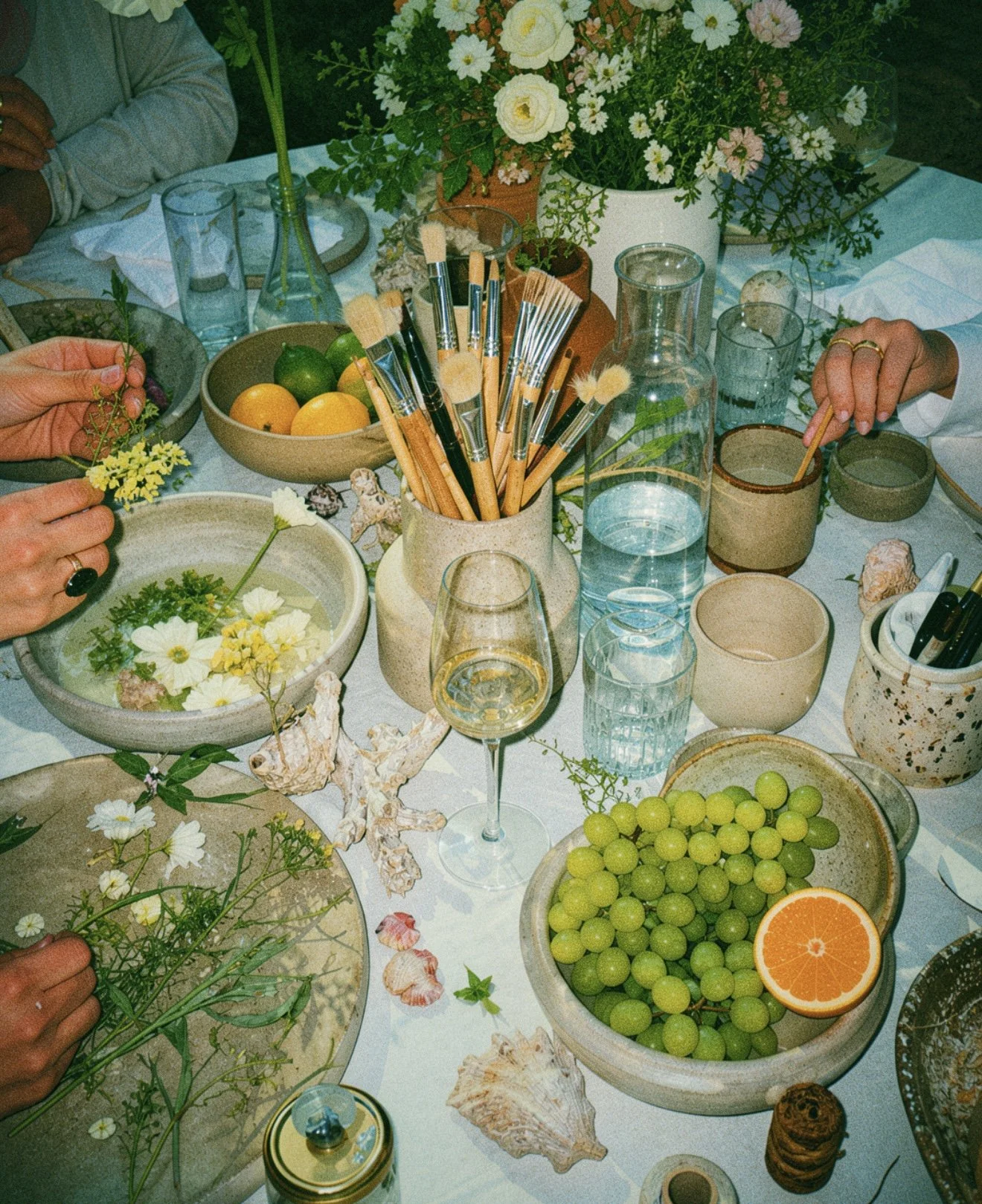 A community wellness creative event. A table decorated with a large floral centerpiece, fresh fruit including grapes and an orange, glassware, and various small containers. People are arranging flowers and participating in a creative activity.