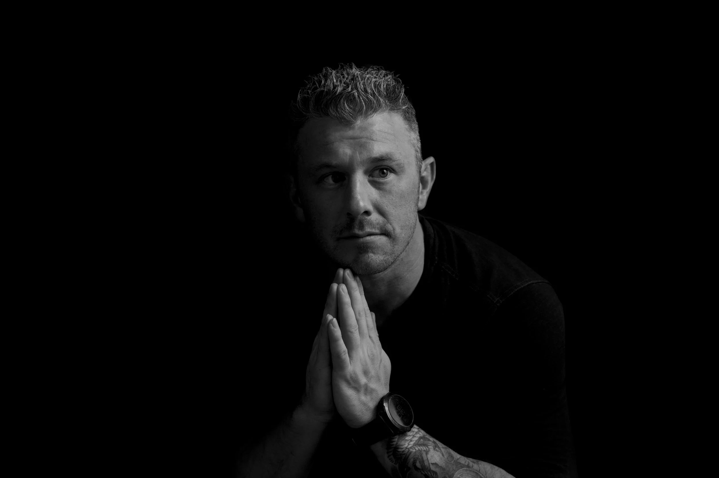 Black and white portrait of a man with curly hair, clasping his hands together and looking upwards, with a dark background.