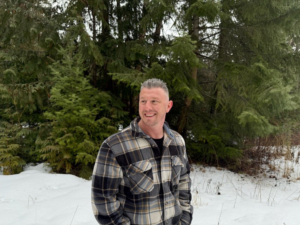 A man in a plaid flannel shirt standing outdoors in a snowy forest, smiling and looking to his left.