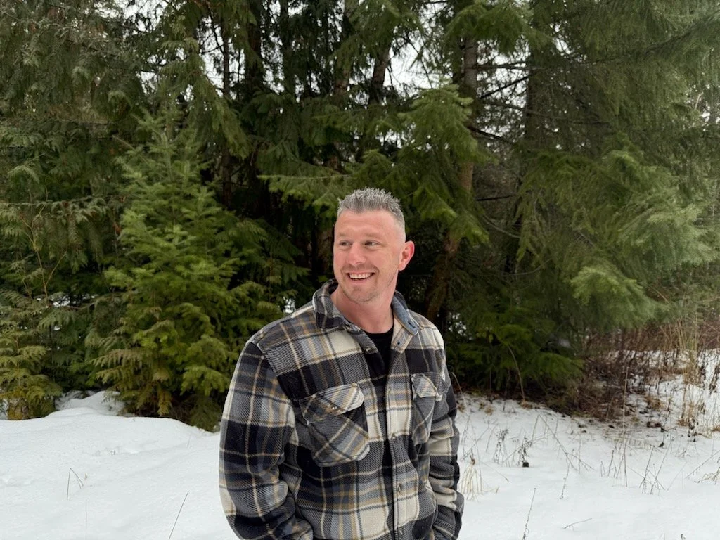 A man standing outdoors in a snowy forested area, wearing a plaid jacket and smiling.