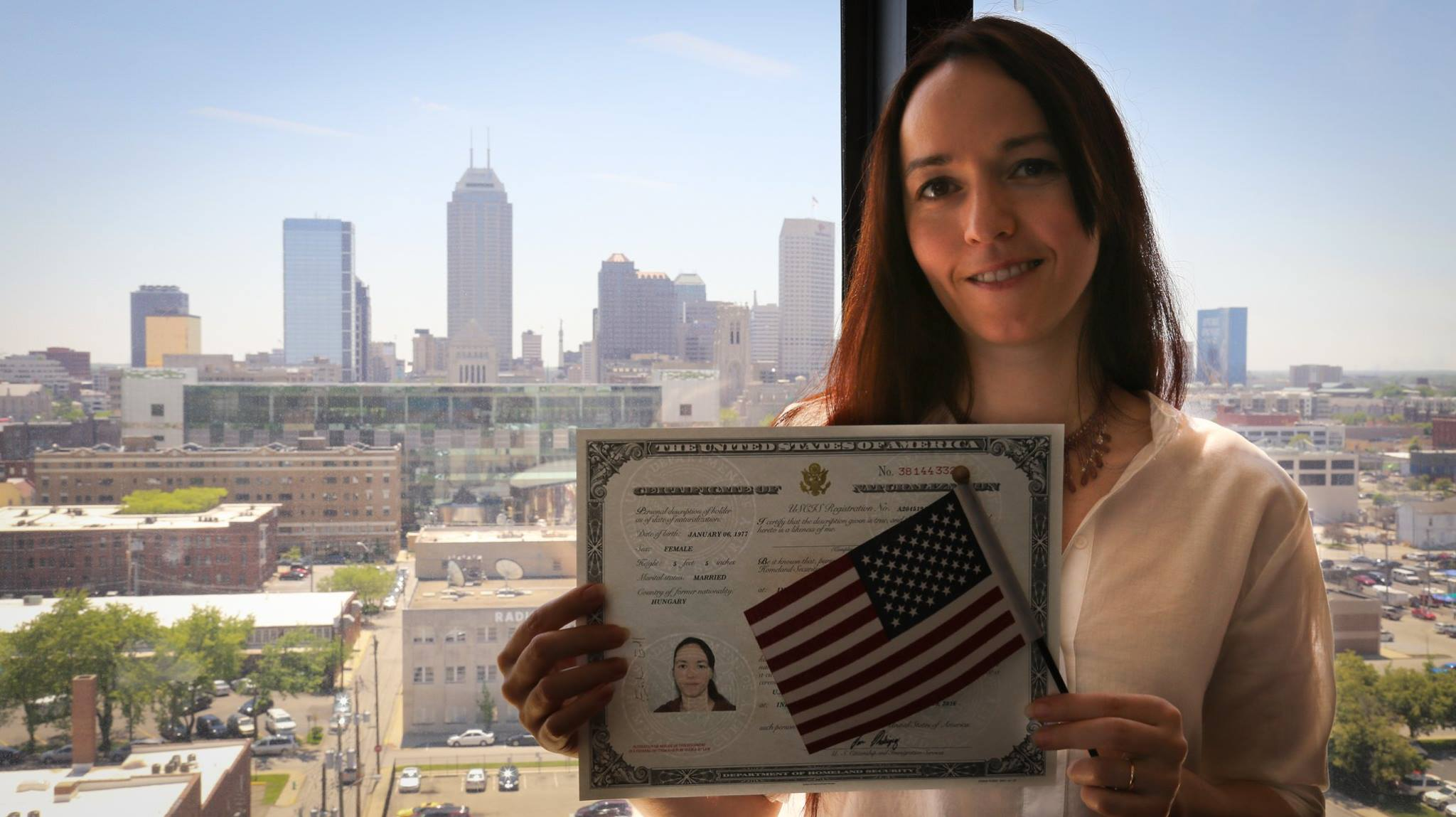 Woman in a beige blazer holding a U.S. citizenship certificate, small American flags, and smiling in front of a city skyline with tall buildings on a sunny day.