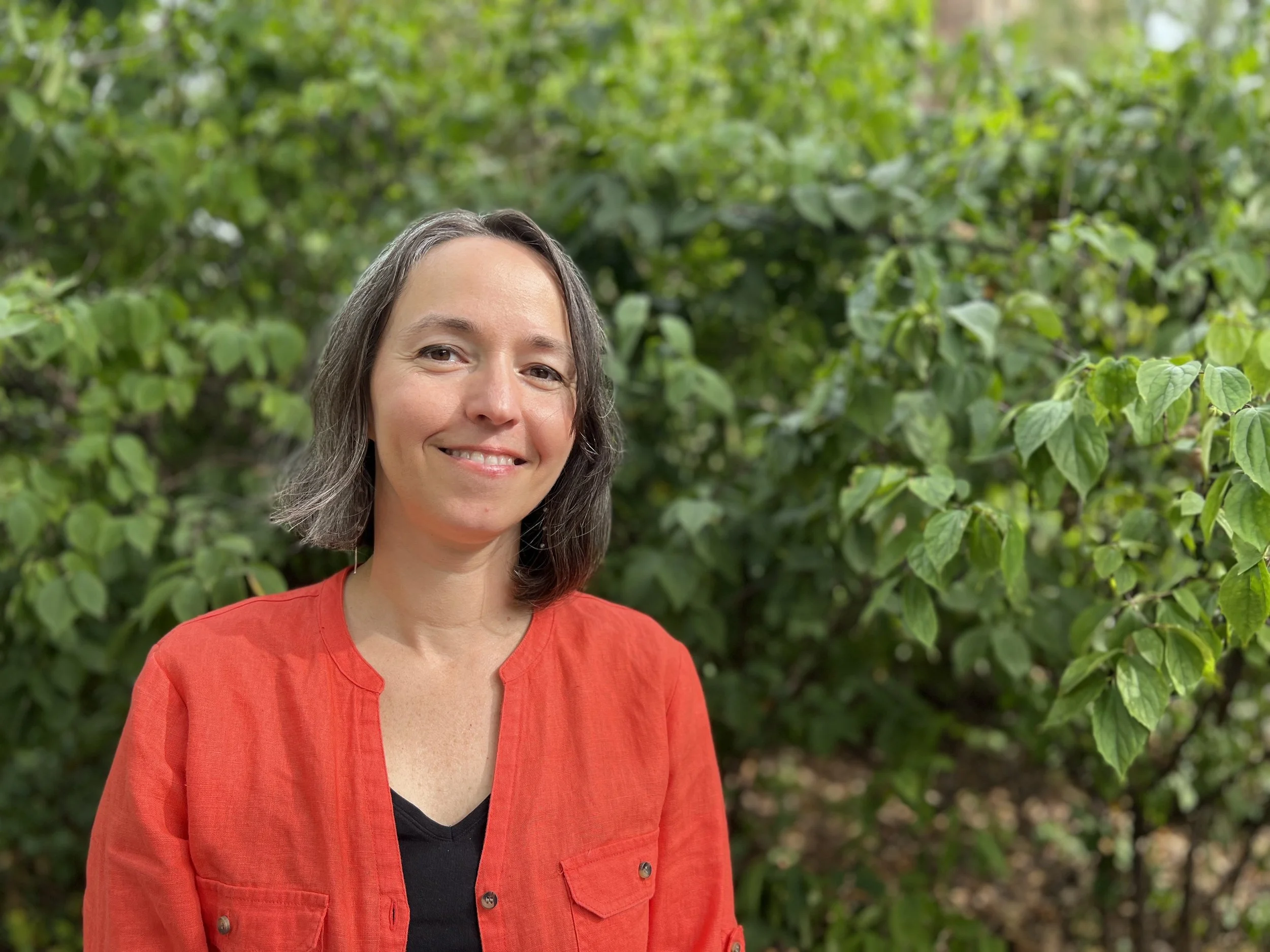 A woman with shoulder-length gray hair smiling, wearing a red jacket over a black shirt, standing outdoors in front of green foliage.