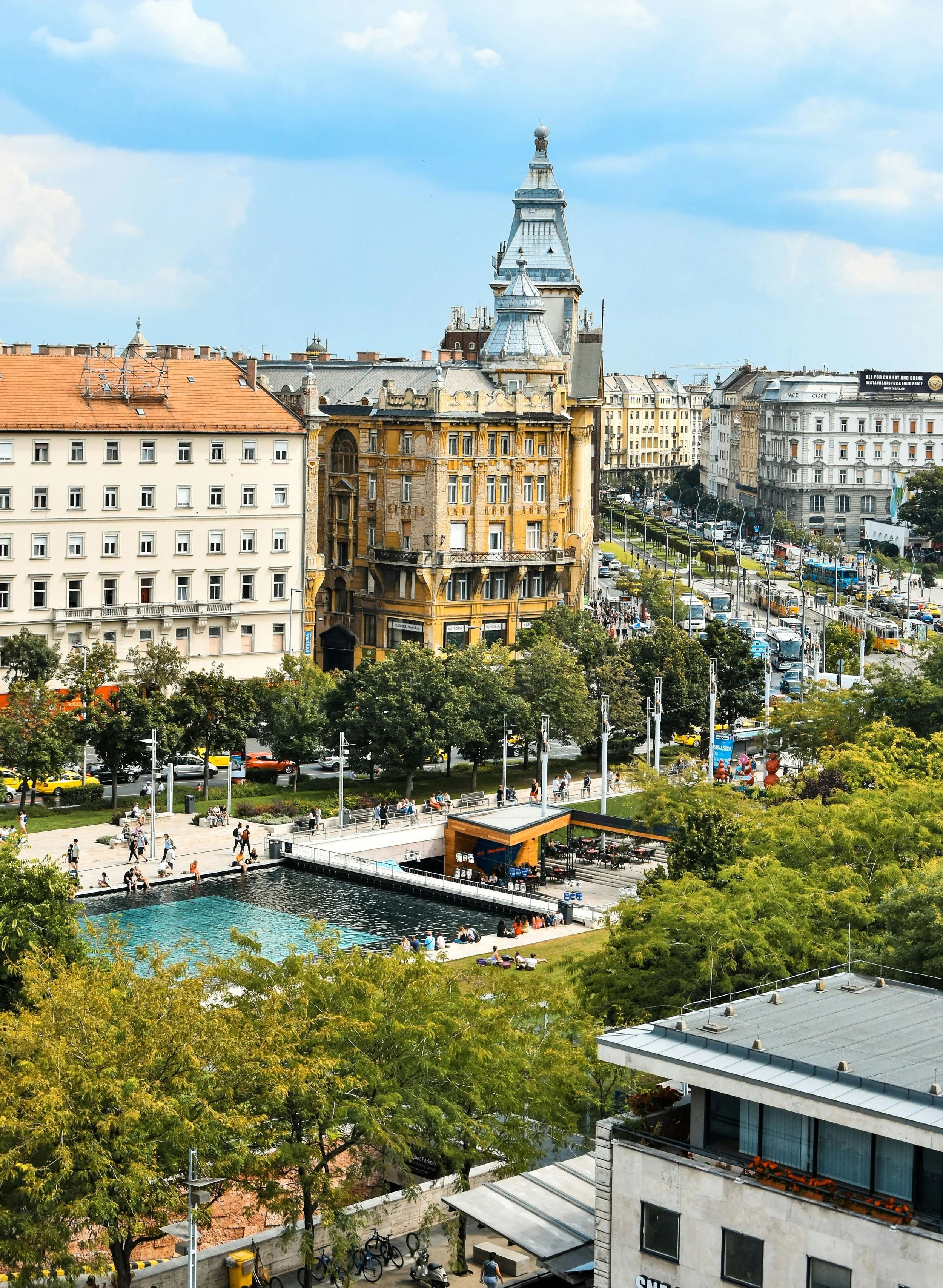 Cityscape of Budapest with historic building, busy street, and park with people, trees, and a water feature