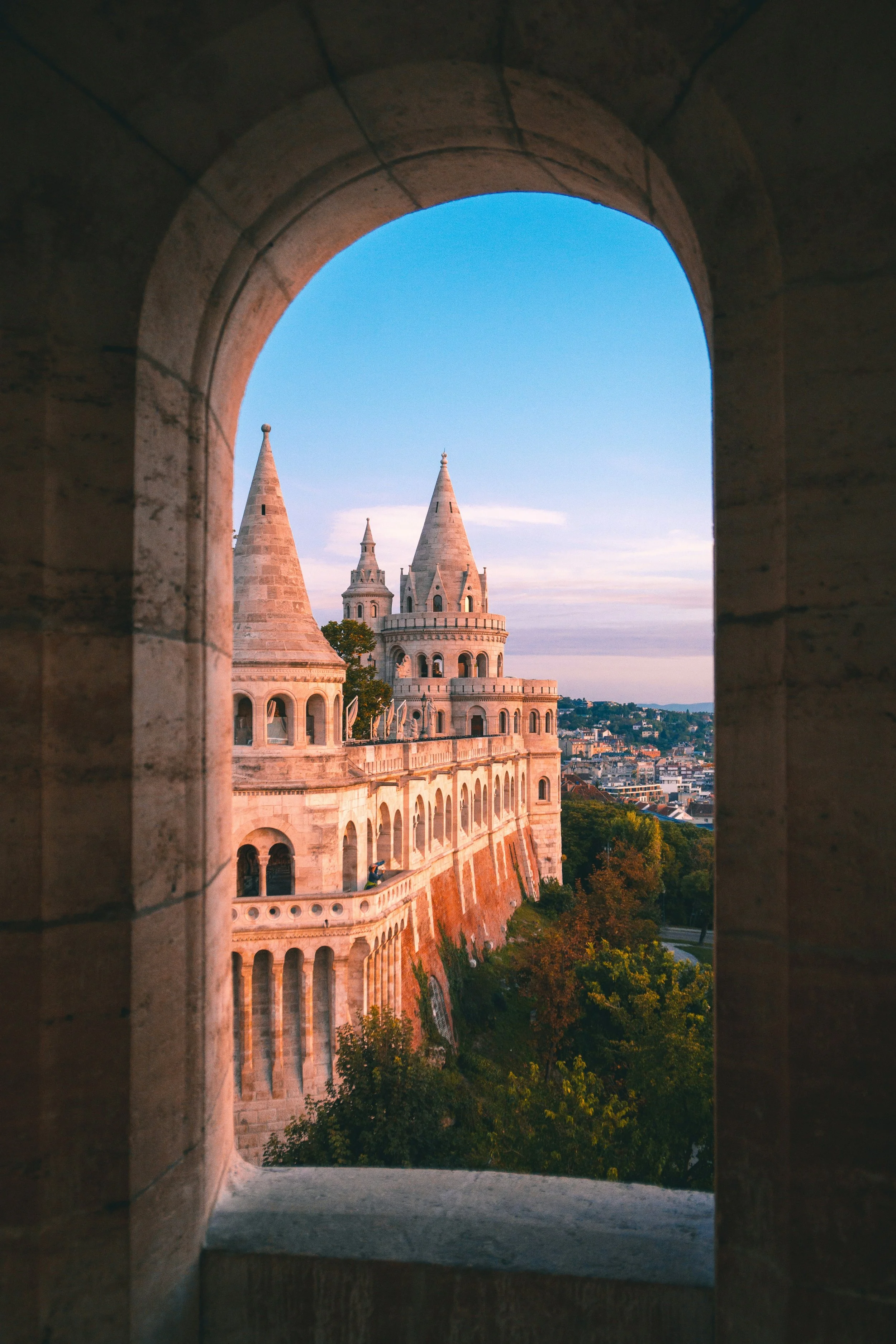 View of Fisherman's Bastion in Budapest, Hungary, seen through a stone arch window at sunset, with the city and trees in the background.