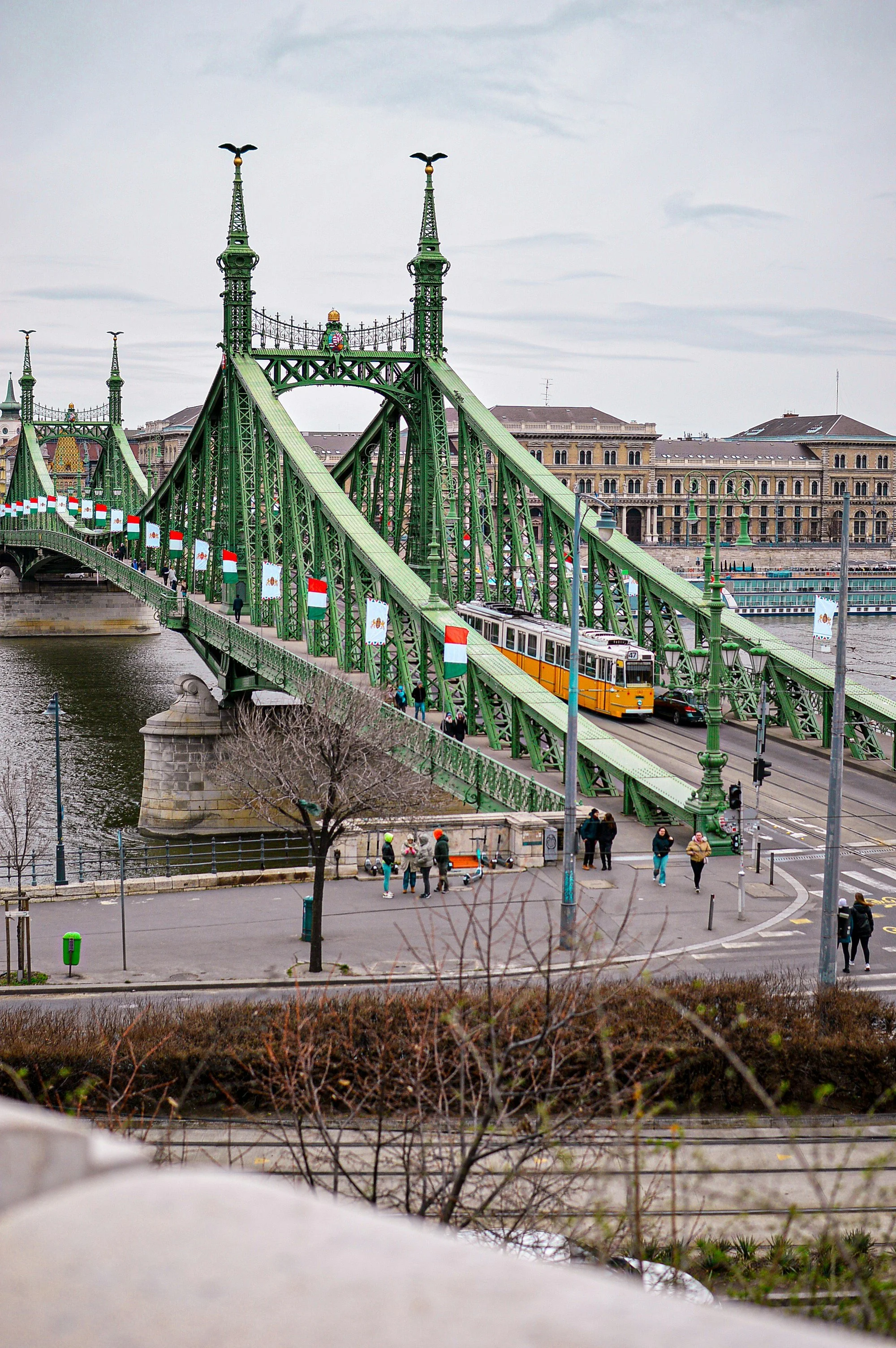 View of the Freedom bridge in Budapest with a tram crossing over a river, with historic buildings and a cloudy sky in the background. People are walking on the sidewalk.