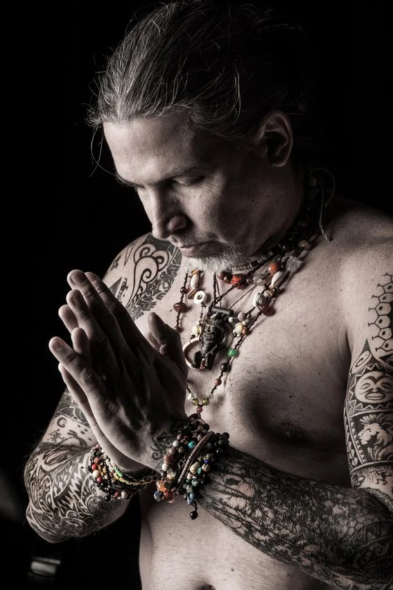 A shirtless man with long gray hair and numerous tattoos on his arms and chest, wearing multiple beaded necklaces and bracelets, holds his hands together in a prayer-like gesture with his eyes closed.