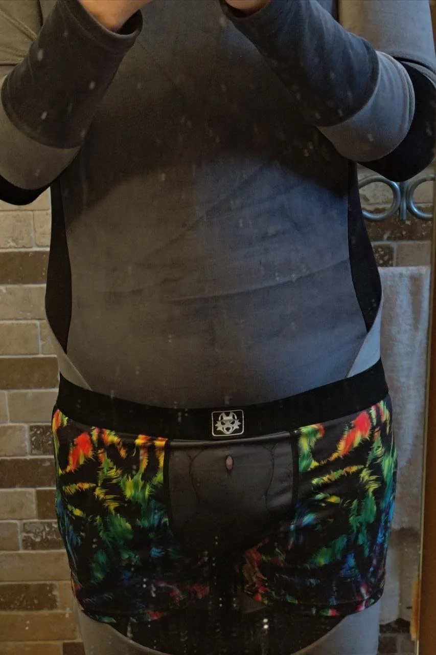 Person taking a mirror selfie wearing a gray and black long sleeve shirt and colorful shorts with a black waistband featuring a wolf logo, standing in a bathroom with a brick wall background.