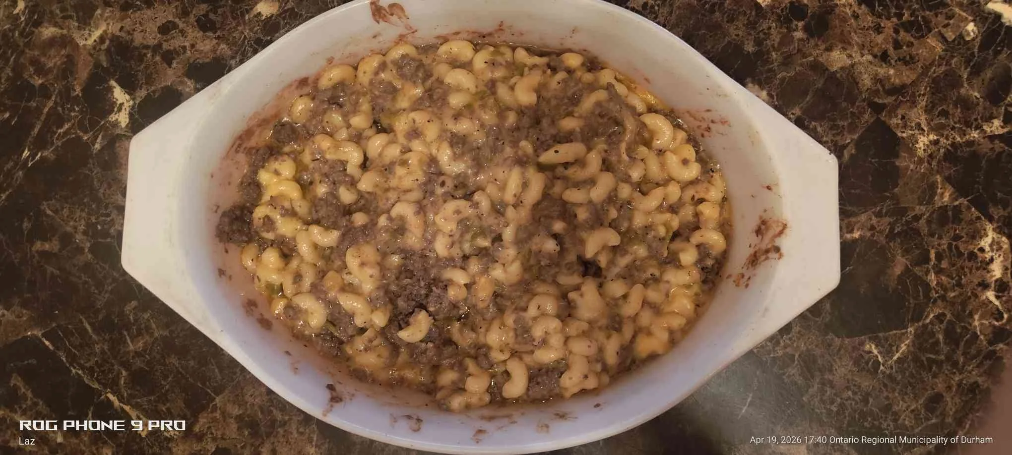A white bowl filled with cooked macaroni and ground beef, placed on a dark marble countertop.