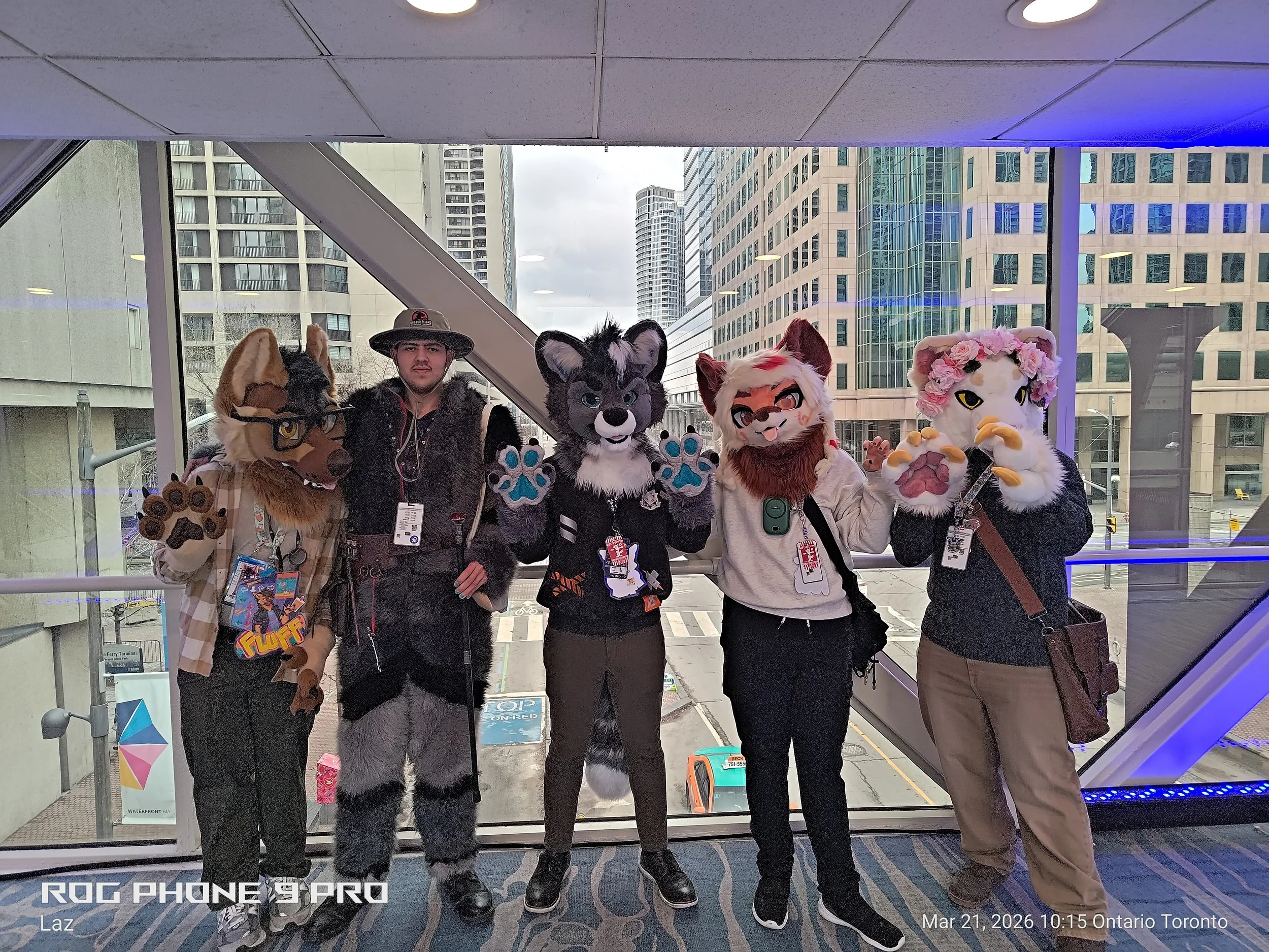 Four people dressed in animal costumes and a person in casual clothing standing inside a glass-enclosed walkway with city buildings in the background.