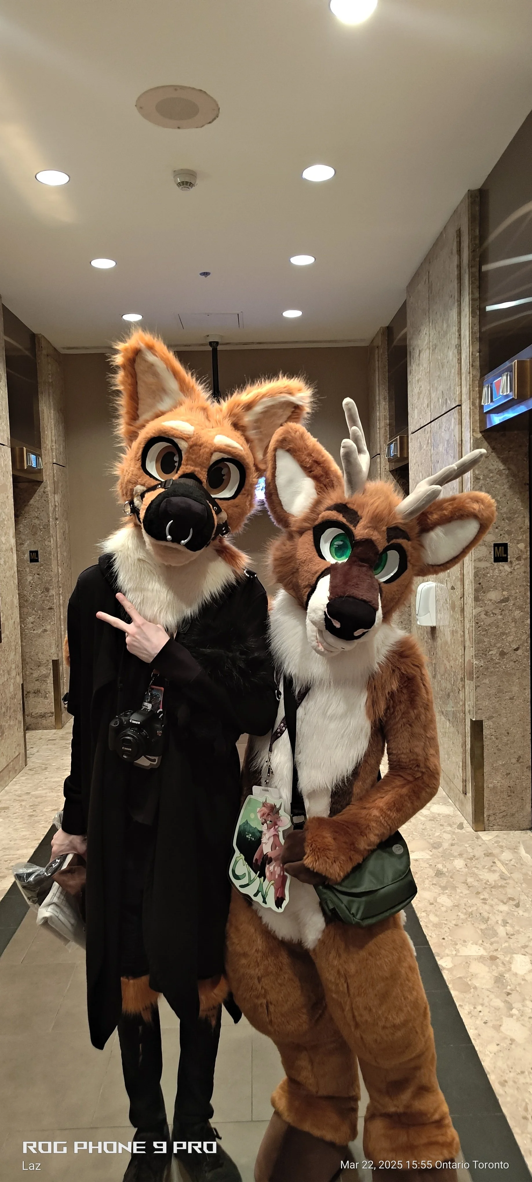 Two people in animal costumes, a fox and a deer, standing in an indoor hallway, smiling at the camera. The person dressed as a fox has a large head with orange fur, big eyes, and a black nose, wearing a black jacket and holding a camera and some item