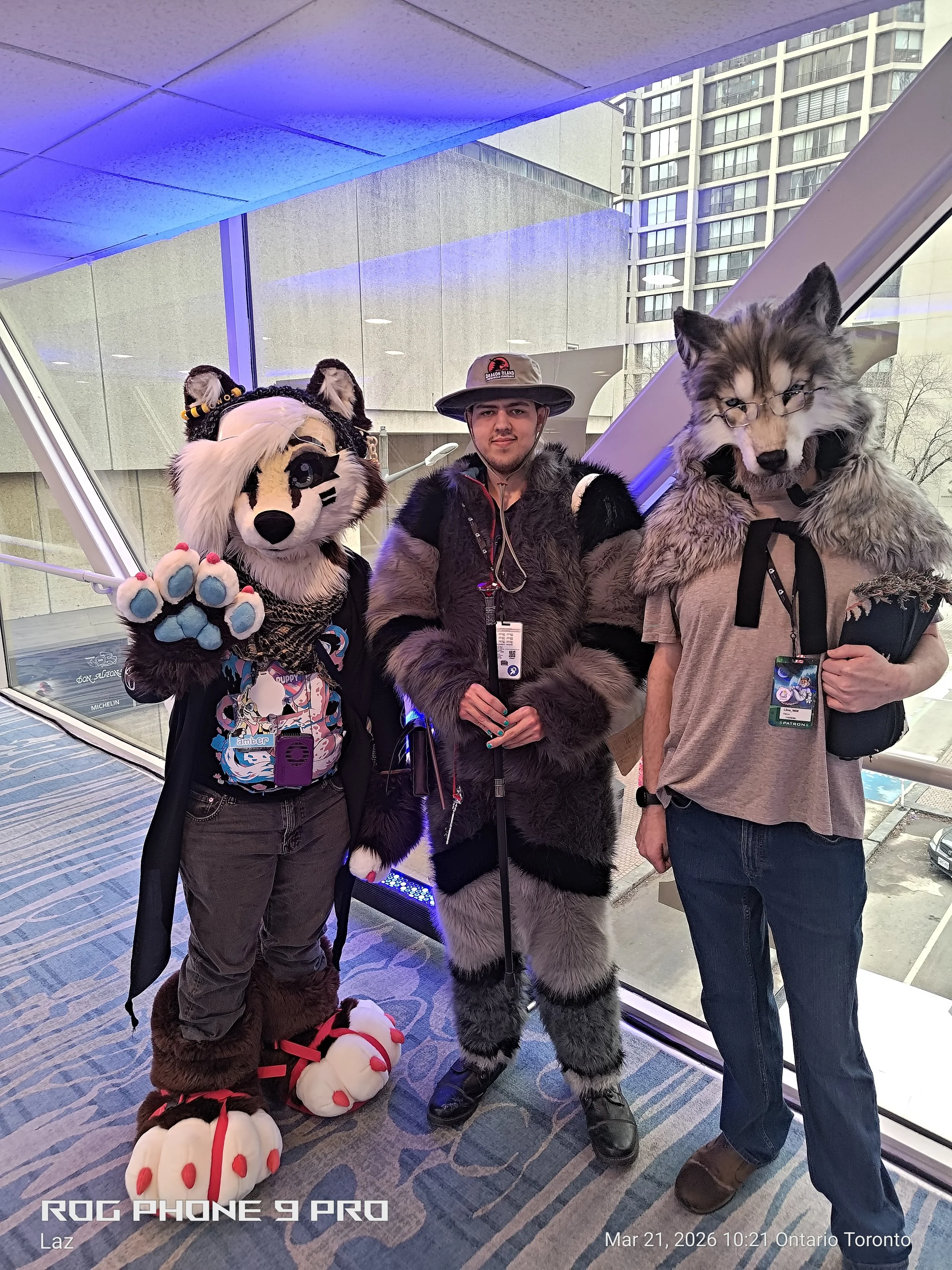 Three people in costumes standing inside a building near large windows, with an urban cityscape visible outside. The person on the left is dressed as a dog with a furry costume, the middle person is dressed as a wolf in a furry suit and glasses, and 