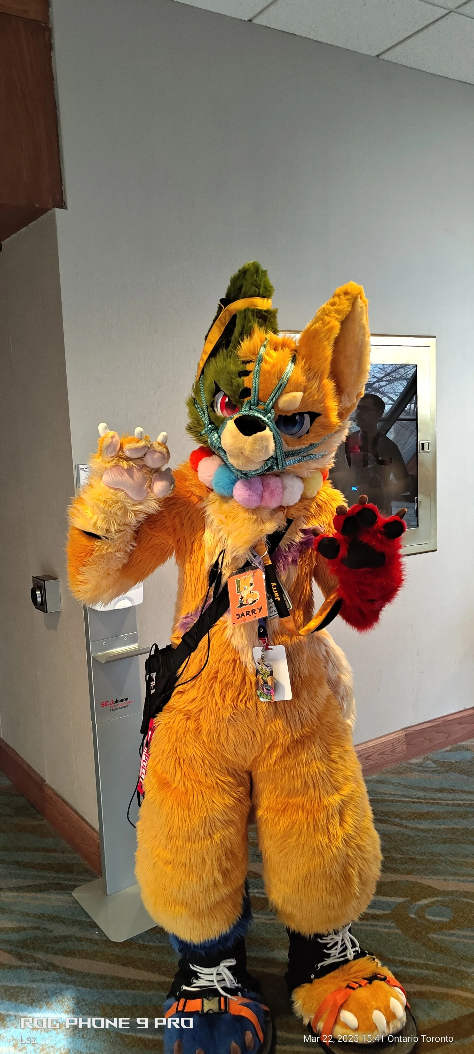 Person in a colorful, furry fox costume with a multicolored plush collar and paw gloves, posing indoors at an event.