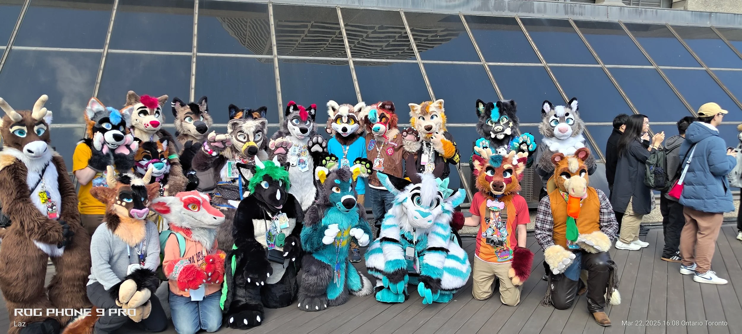 Group of people in colorful animal costumes, known as furries, gathered outdoors in front of a modern glass building, with some taking photos.