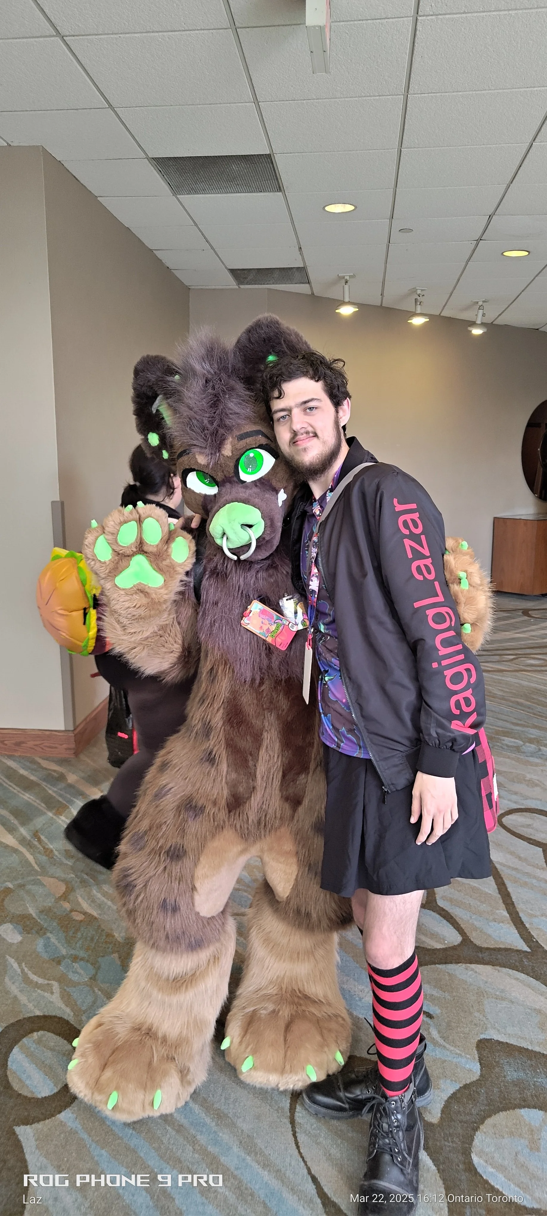 A young man with a beard wearing a black jacket with pink text, a colorful shirt, black shorts, pink and black striped socks, and black shoes standing next to a person in a furry lion costume, inside a convention center or hotel with patterned carpet