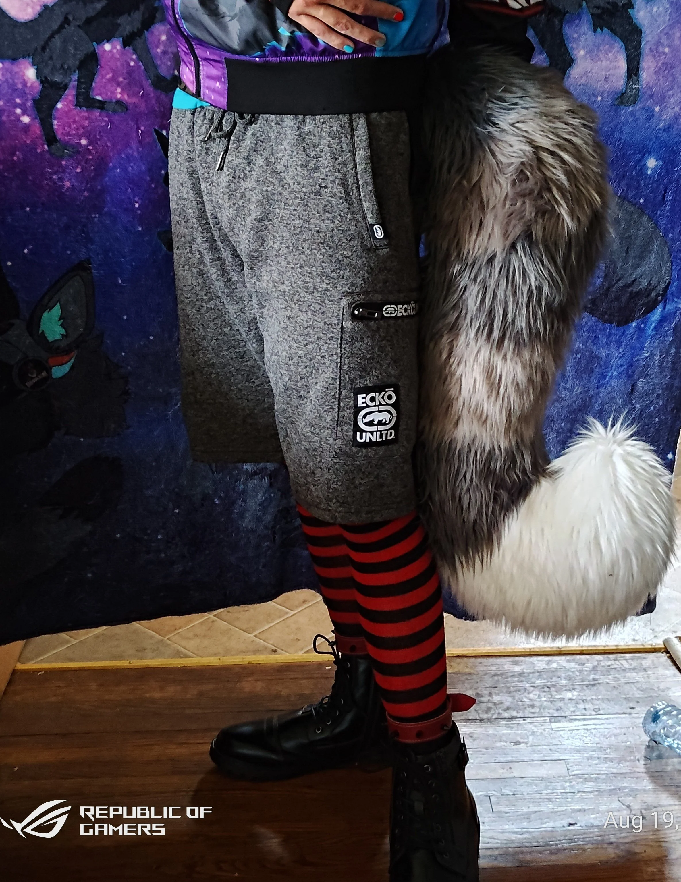 A person wearing black combat boots, red and black striped leggings, and a gray jacket with patches, standing on a wooden floor in front of a dark, space-themed backdrop with unicorns. The person's hand, with painted nails, is resting on their chest.