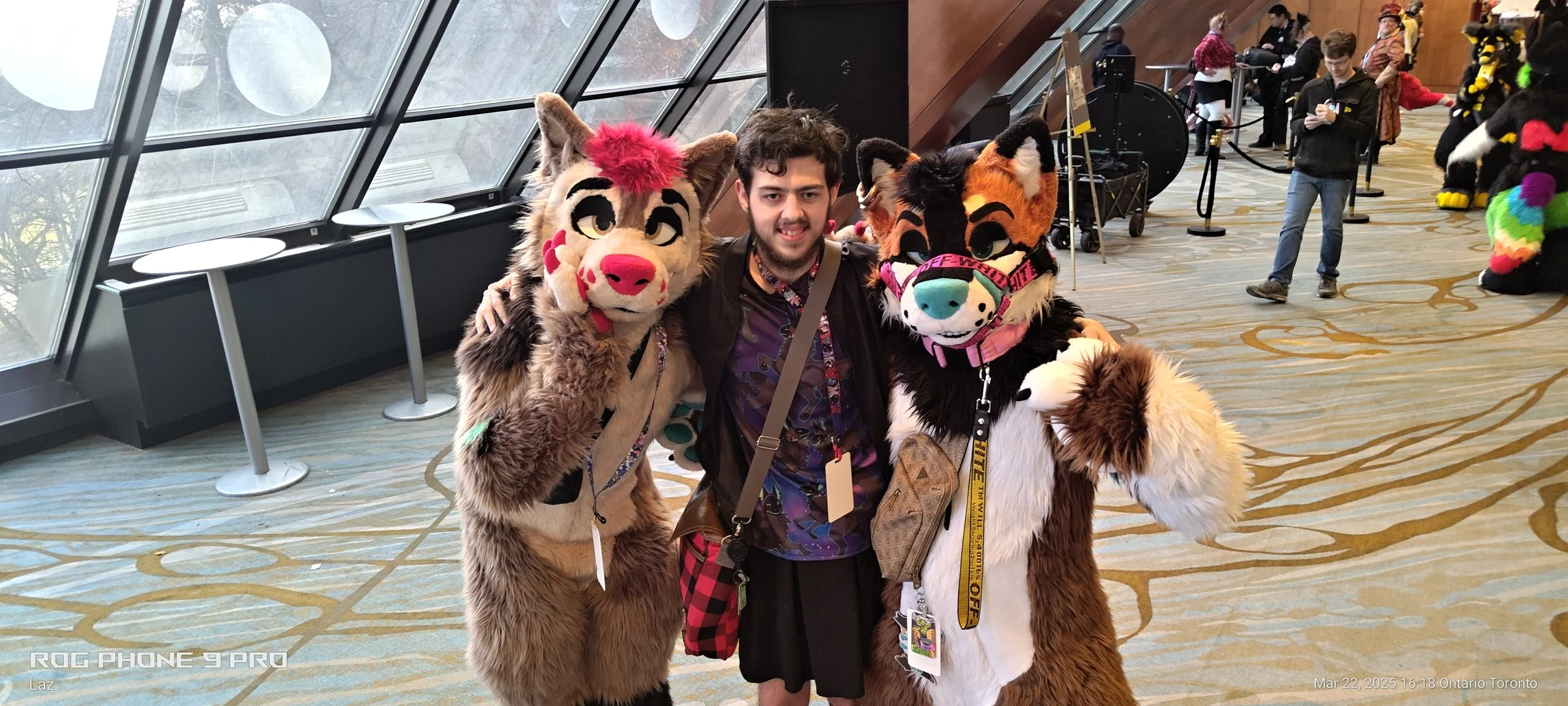 A man with two furry mascot costumes, one resembling a lion and the other an anthropomorphic animal with glasses, posing together indoors at a convention or event.