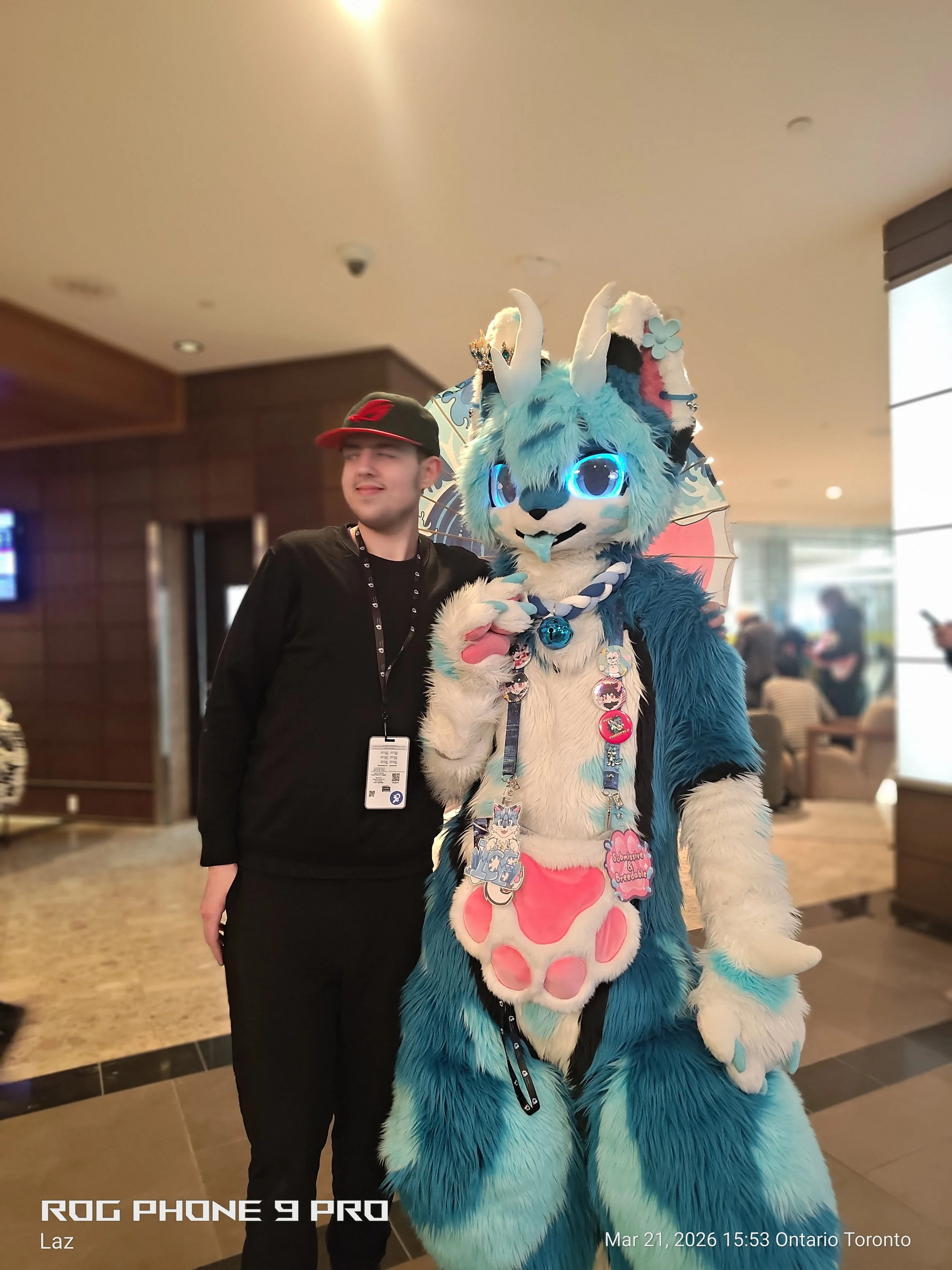 A person dressed in a colorful, furry dragon or mascot costume with large glowing eyes, standing next to a person in casual clothing in an indoor setting, with other people in the background.