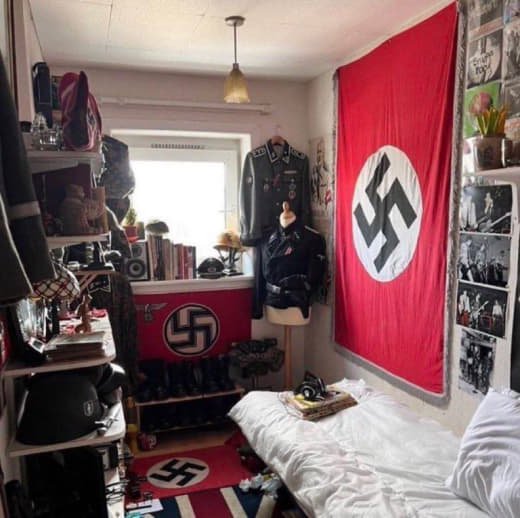 A bedroom decorated with Nazi symbols, including a large swastika flag on the wall and a smaller swastika banner on a shelf. The room contains military-style uniforms, books, and photos, with a bed in the corner and various personal items.