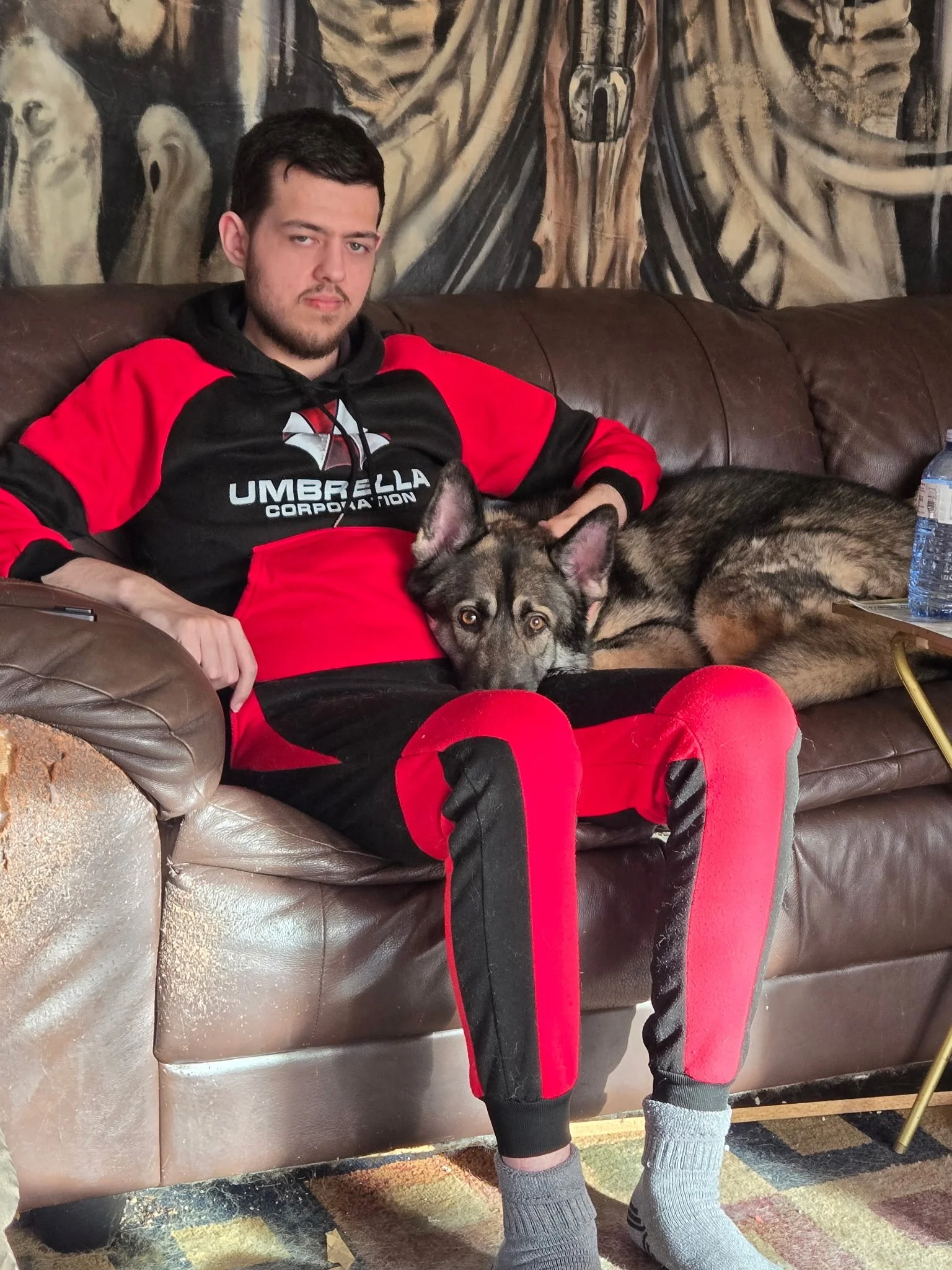 A young man wearing a black and red hoodie with the Umbrella Corporation logo sitting on a brown leather couch. He has dark hair, a beard, and is sticking his tongue out slightly. A dog with a black, gray, and tan coat is lying next to him, resting i