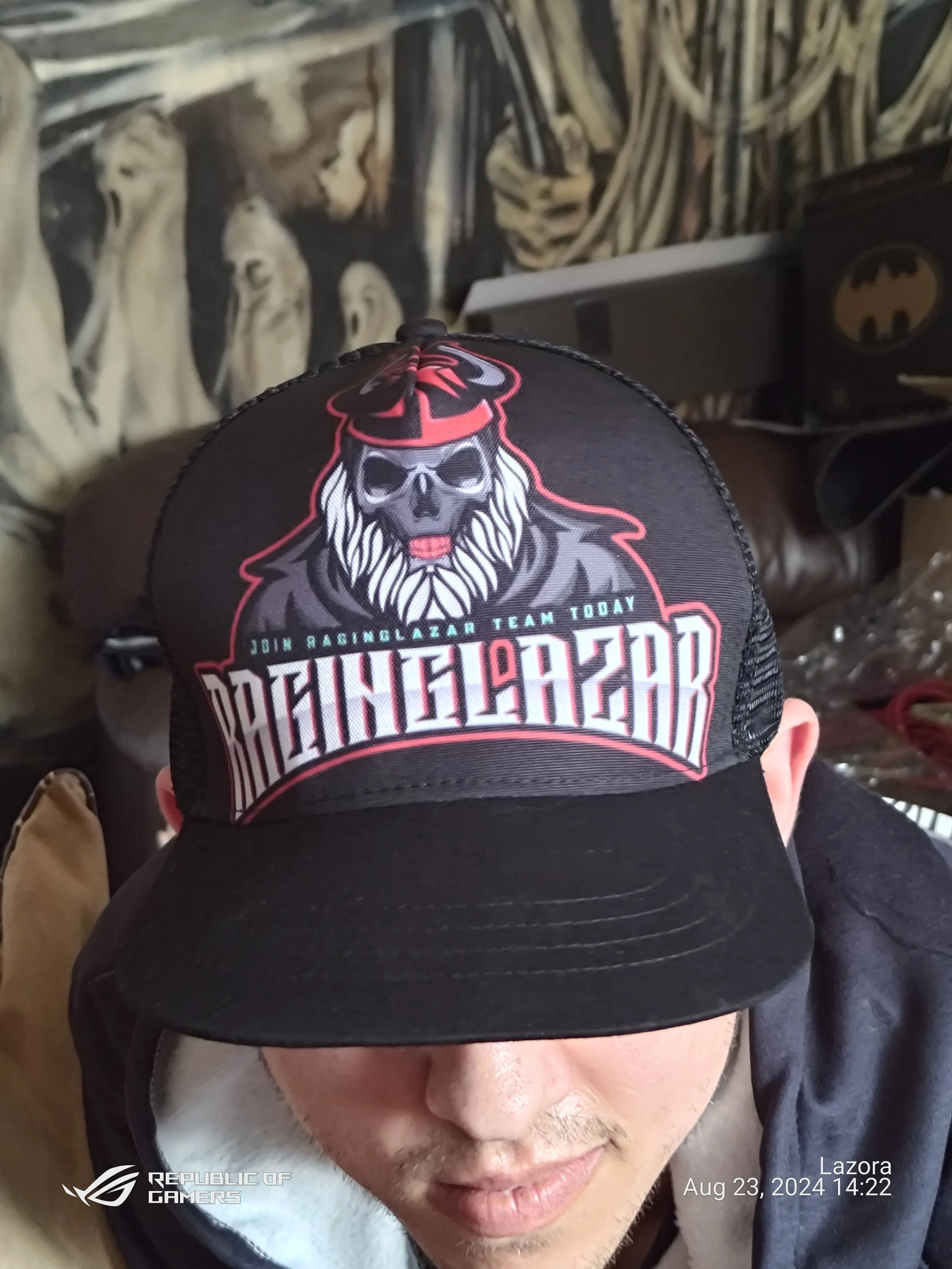 A person wearing a black cap with a skull, lion, and Batman logo design, partially covering their face, with a background featuring wall art of skulls and images of skeletons.