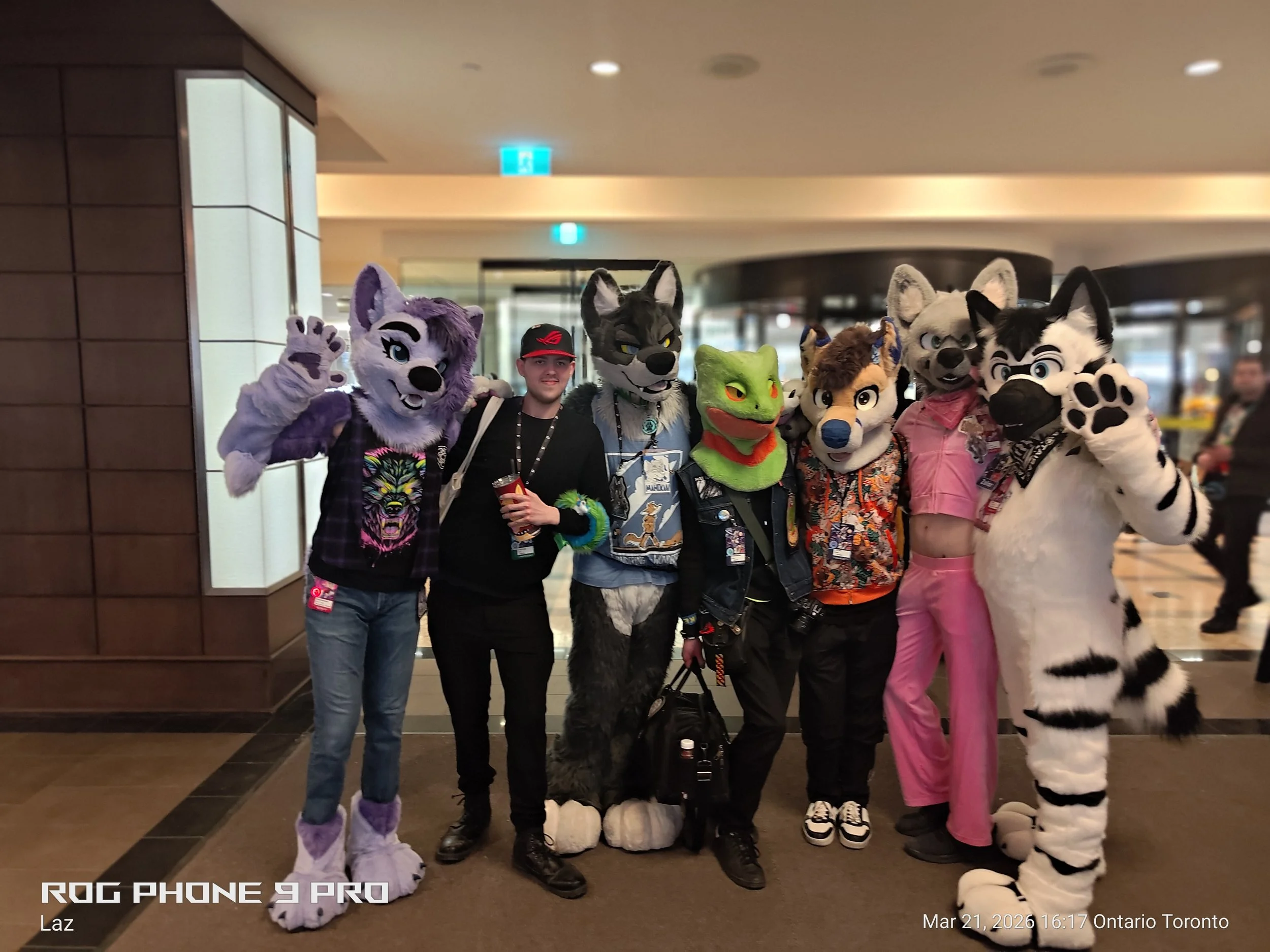 Group of seven people, some in furry animal costumes, standing together in a lobby or convention center. The costumes include various animal characters such as wolves, a frog, and a tiger. The others are wearing casual clothing and lanyards.