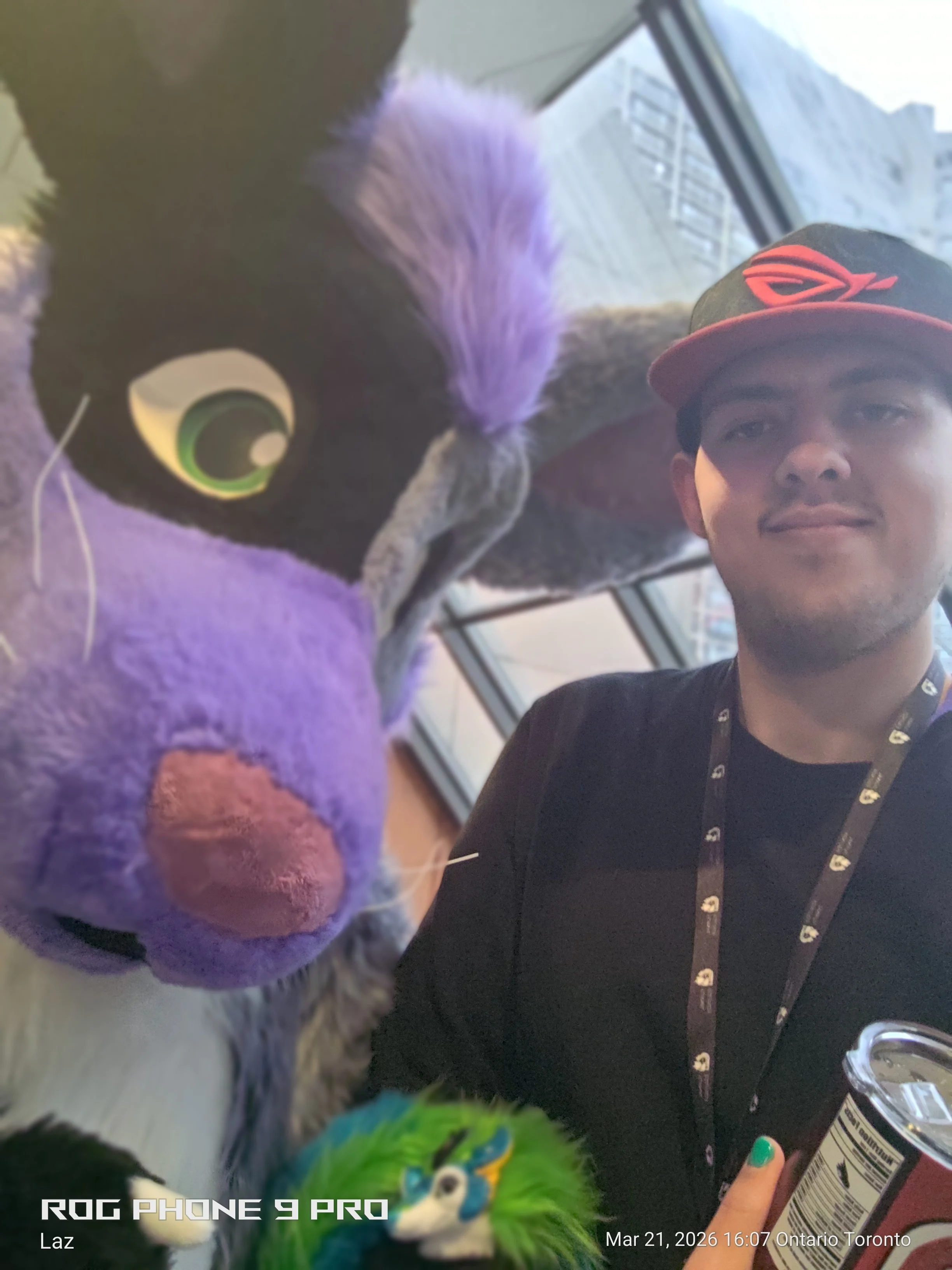 A person wearing a black shirt and a red and black cap taking a selfie with a colorful plush toy resembling a bird with blue, green, and black feathers, purple flower accessories, and a large eye. The background shows a window with a cityscape view a