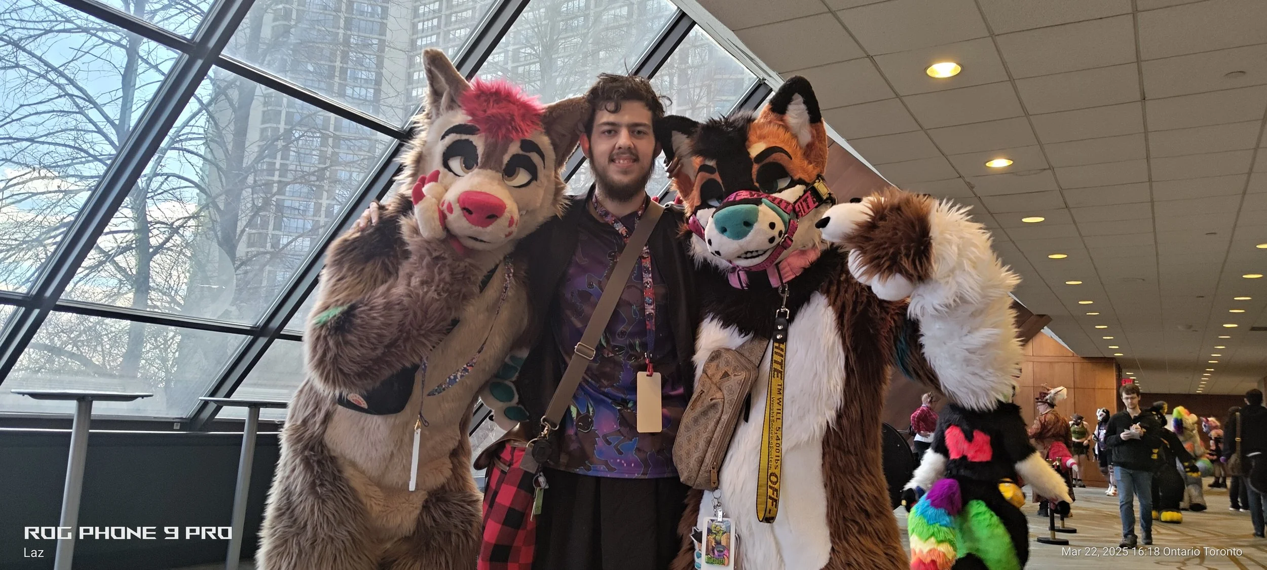 A person in a black jacket and colorful shirt posing with three individuals in furry costumes at a convention center with large windows and other attendees in the background.