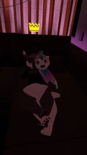 A cartoon unicorn plush toy sitting on a bed with a blanket, illuminated by a faint pinkish light, with a yellow paper crown stuck on a headboard in the background.