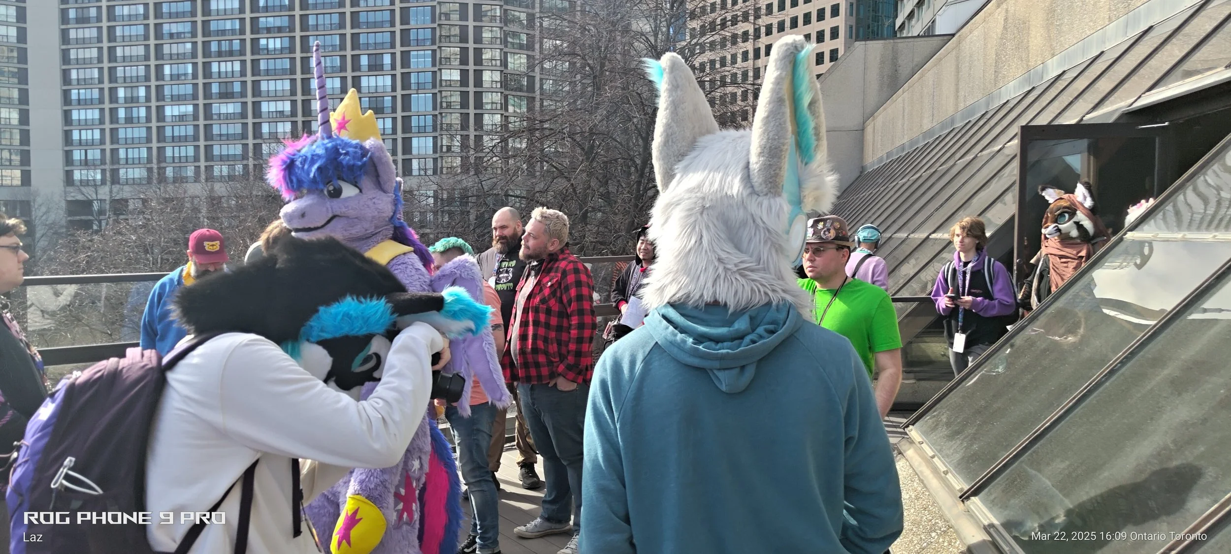 People gathered outdoors in a city, some dressed in furry animal costumes and hoodies, talking and interacting with each other. Tall buildings and trees are in the background.