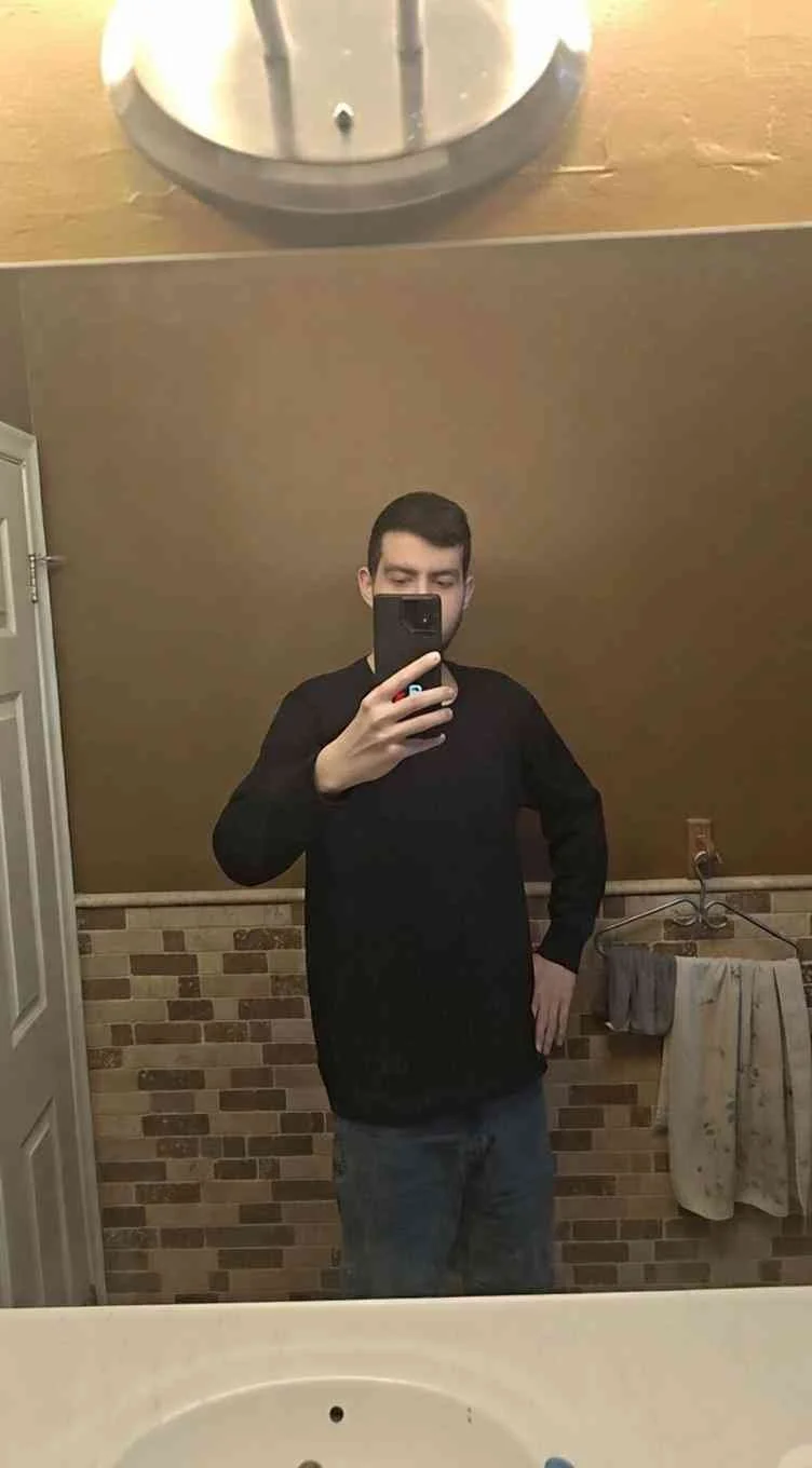A young man is taking a mirror selfie in a bathroom, wearing a black long-sleeve shirt and jeans. The bathroom has a brick-patterned lower wall, a towel hanging on a hook, and a mirror with a metal frame.