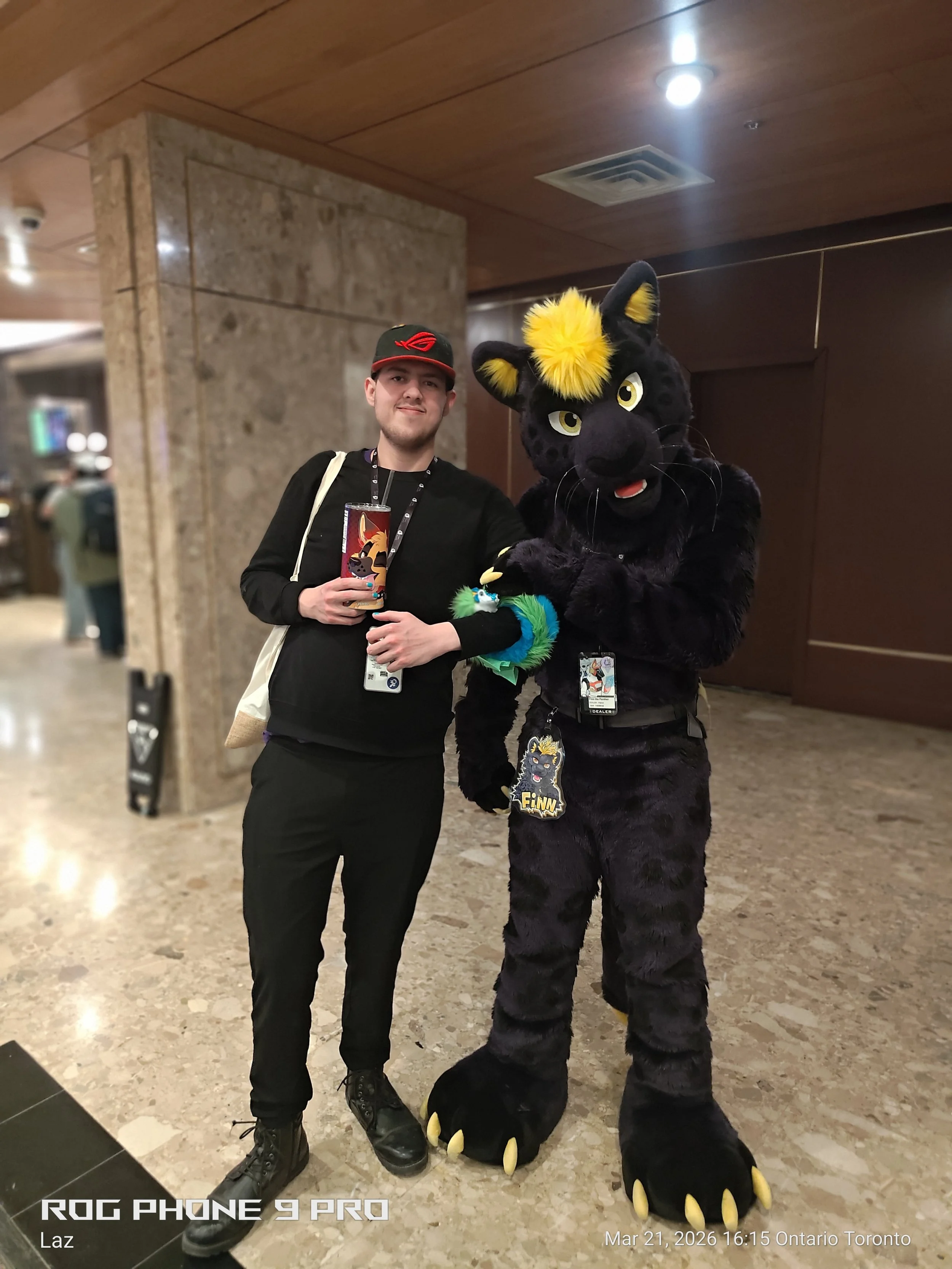 A man dressed in black holding a drink and wearing a hat with a red logo, standing next to a person in a black panther costume with yellow accents on the forehead and paws, in an indoor setting.