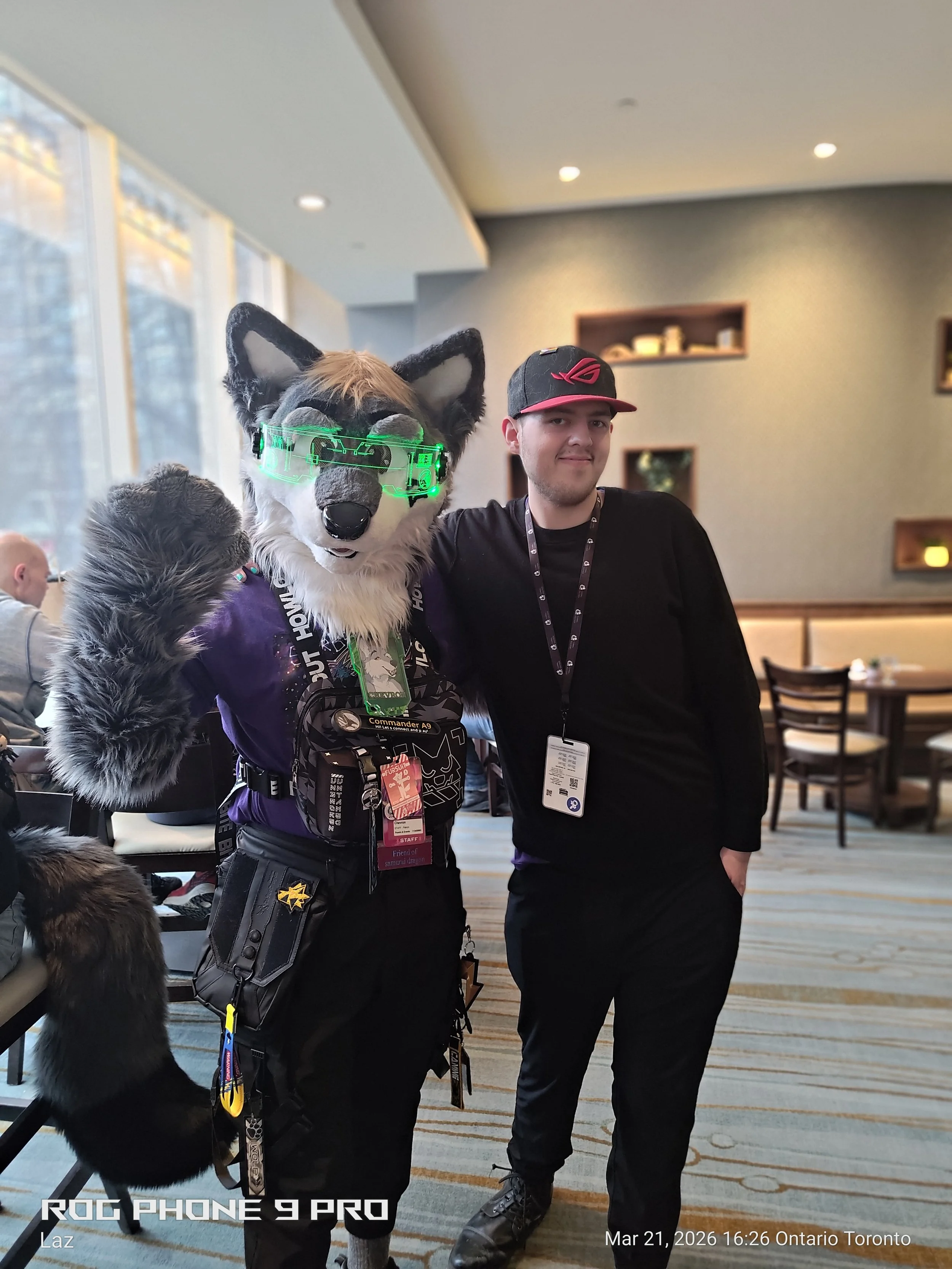 A person dressed in a wolf costume with glowing green goggles standing next to a young man in a black shirt and cap, both posing indoors at a conference or event.