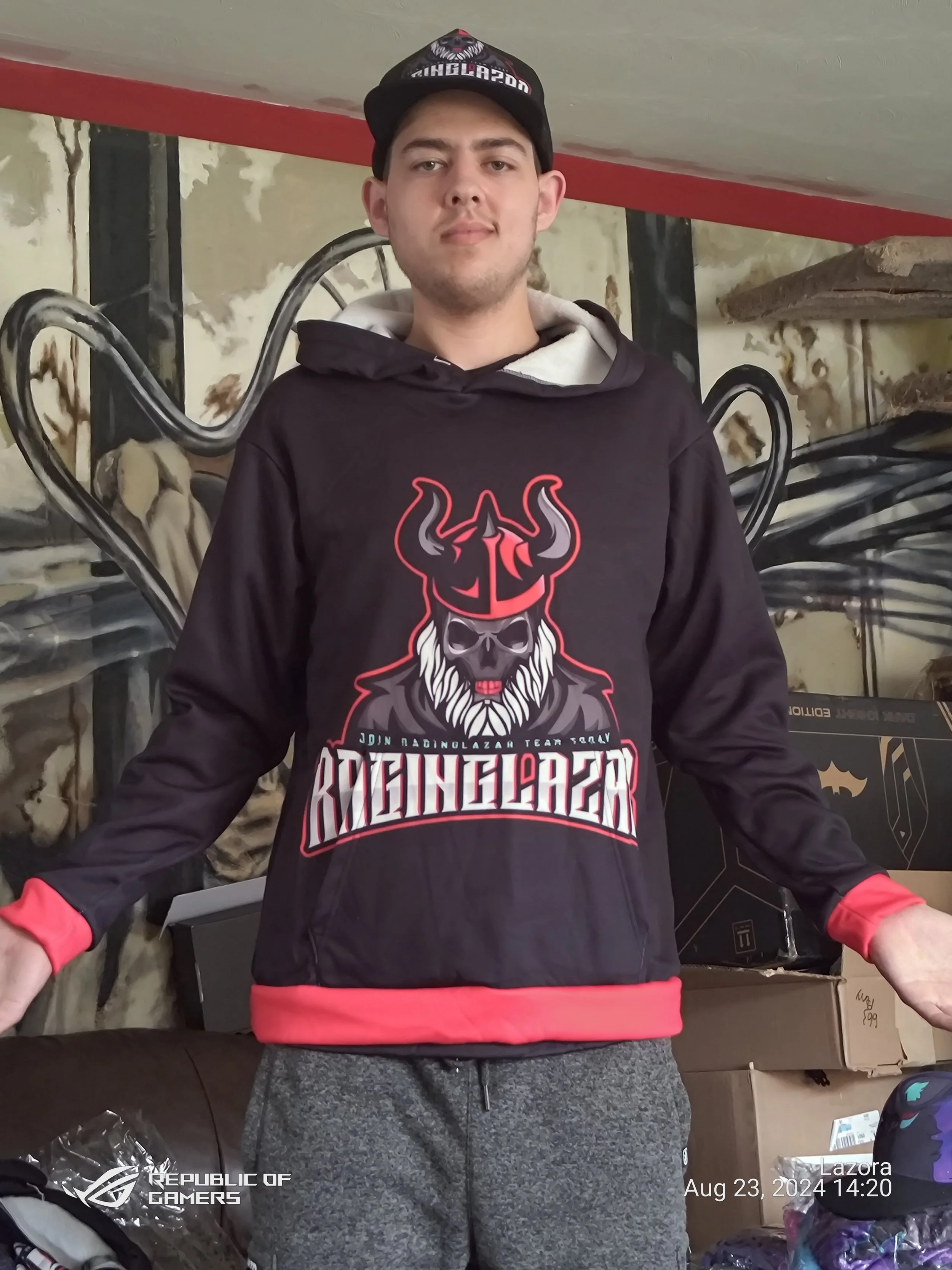 A young man wearing a black hoodie with a graphic design of a skull with a horned helmet, a black cap with a logo, and gray sweatpants standing in a room with boxes and shelves.