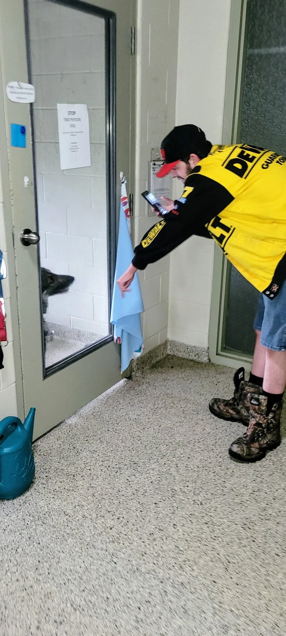 A man wearing a police vest and camouflage boots bending down to feed a black and white dog through a glass door with a sign that says "Stop: No personnel or dog beyond this point."