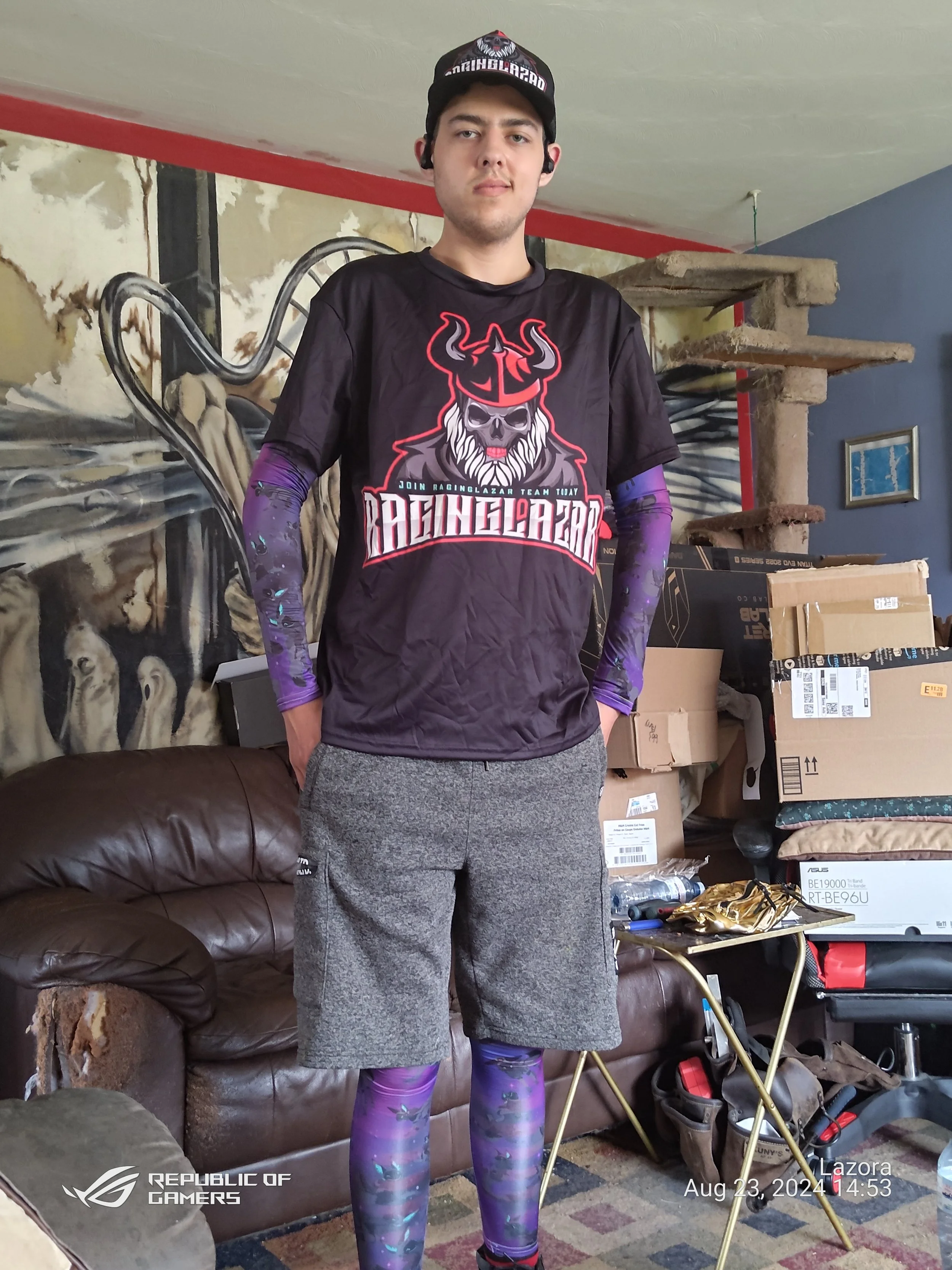 A young man standing indoors in front of a wall with artwork, wearing a black and purple gaming jersey, gray shorts, purple patterned tights, a black cap, and black earbuds.