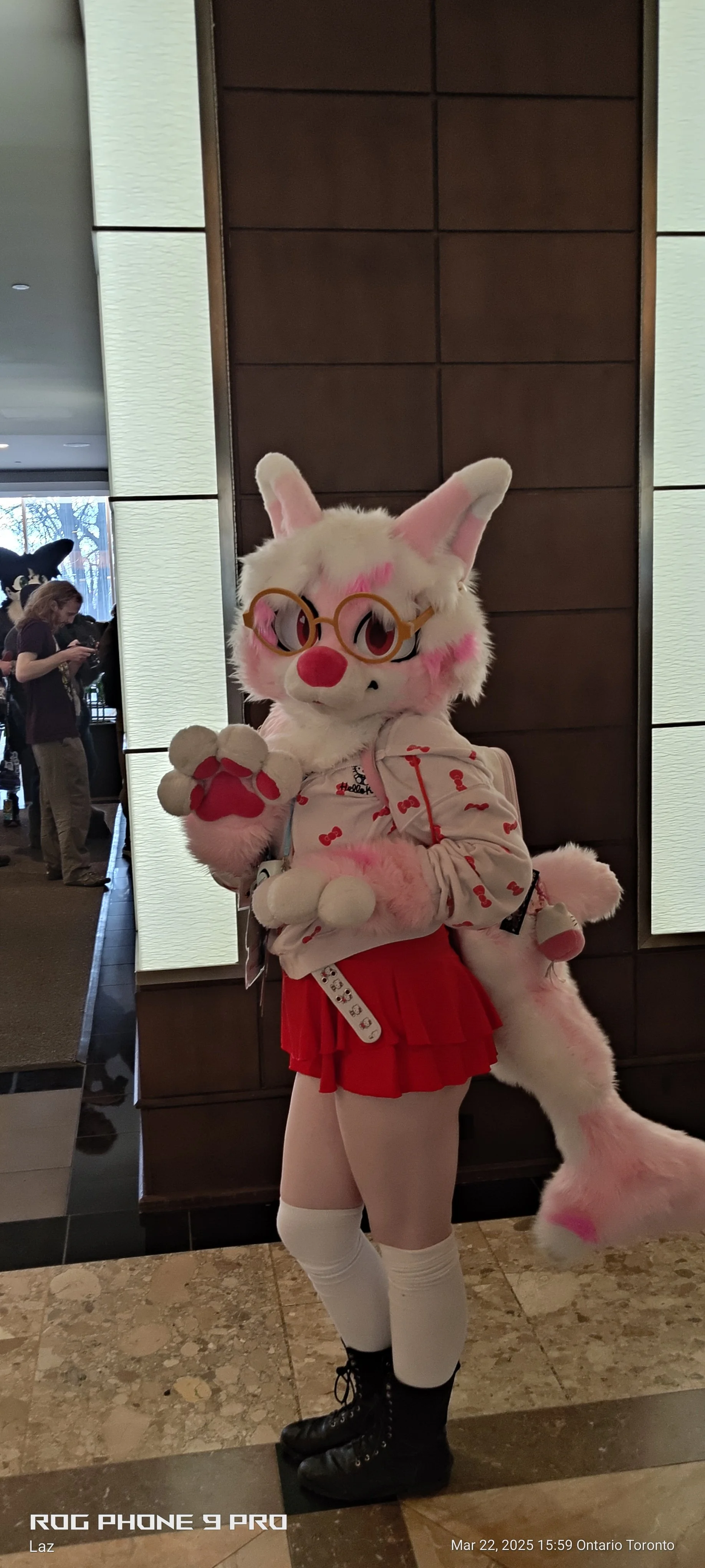 A person in a bunny costume with glasses and a pink and white fluffy tail, dressed in a white and red outfit, at a public event or convention.