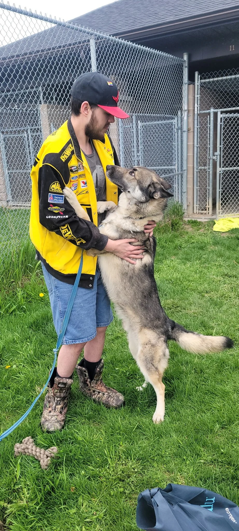 A man with a beard wearing a yellow racing jacket, a black and red cap, blue shorts, and camouflage boots, is standing on grass and hugging a large German Shepherd dog. The dog is standing on its hind legs, leaning against the man, with its front paw