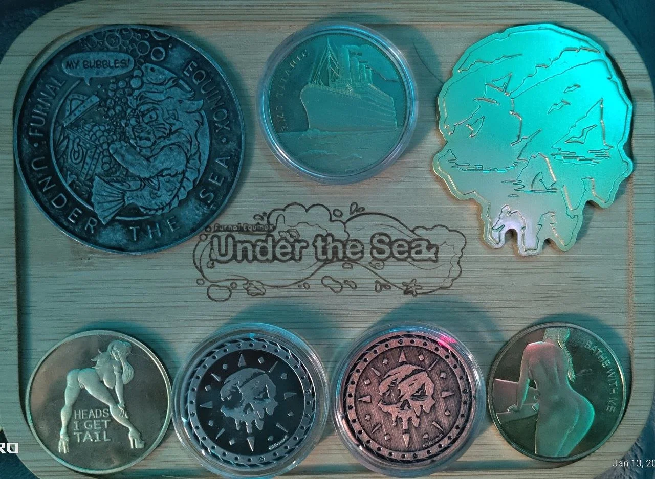 Collection of six tokens on a wooden background, centered around an 'Under the Sea' theme, with one coin featuring a mermaid and the phrase 'Heads I Get Tail,' a coin with a skull, a coin with a ship, a coin with a woman's back, and a coin with a fac