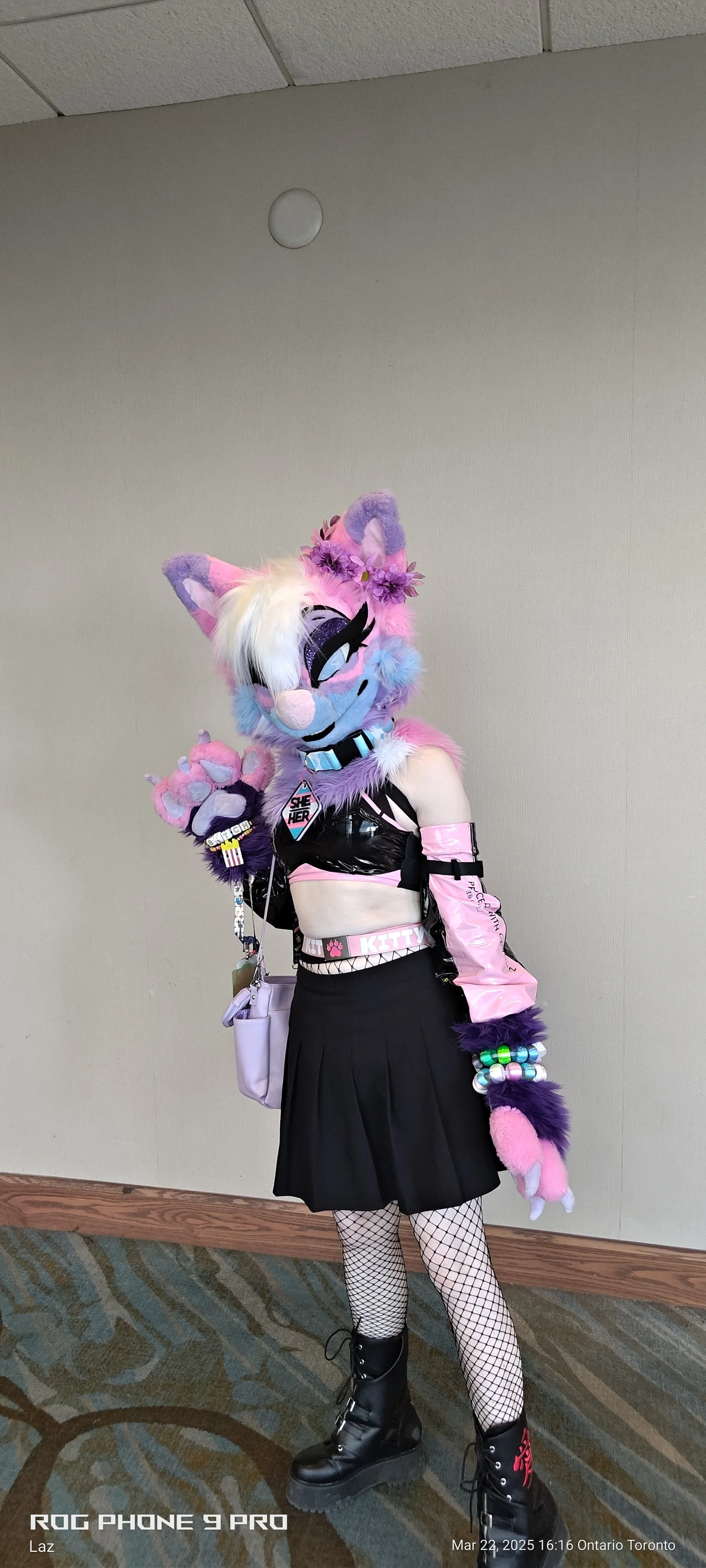 Person dressed in a pink, purple, and black furry cosplay costume with a large animal head mask resembling a pastel-colored, cartoon-style feline character, wearing fishnet stockings, black platform boots, and accessories, standing indoors against a 