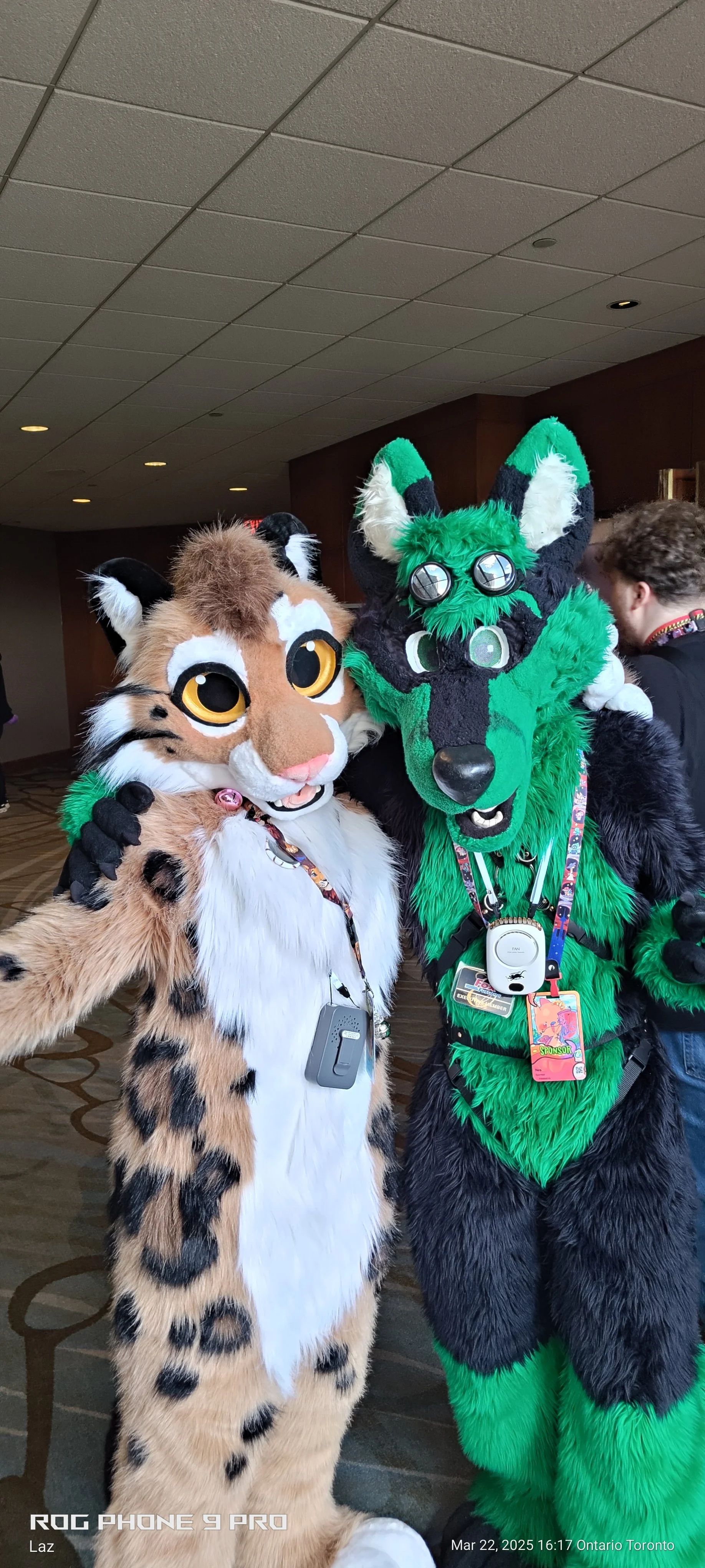 Two people in colorful animal cosplay costumes, one a tan and black spotted feline with large eyes, and the other a green and black wolf or fox, posing together indoors.