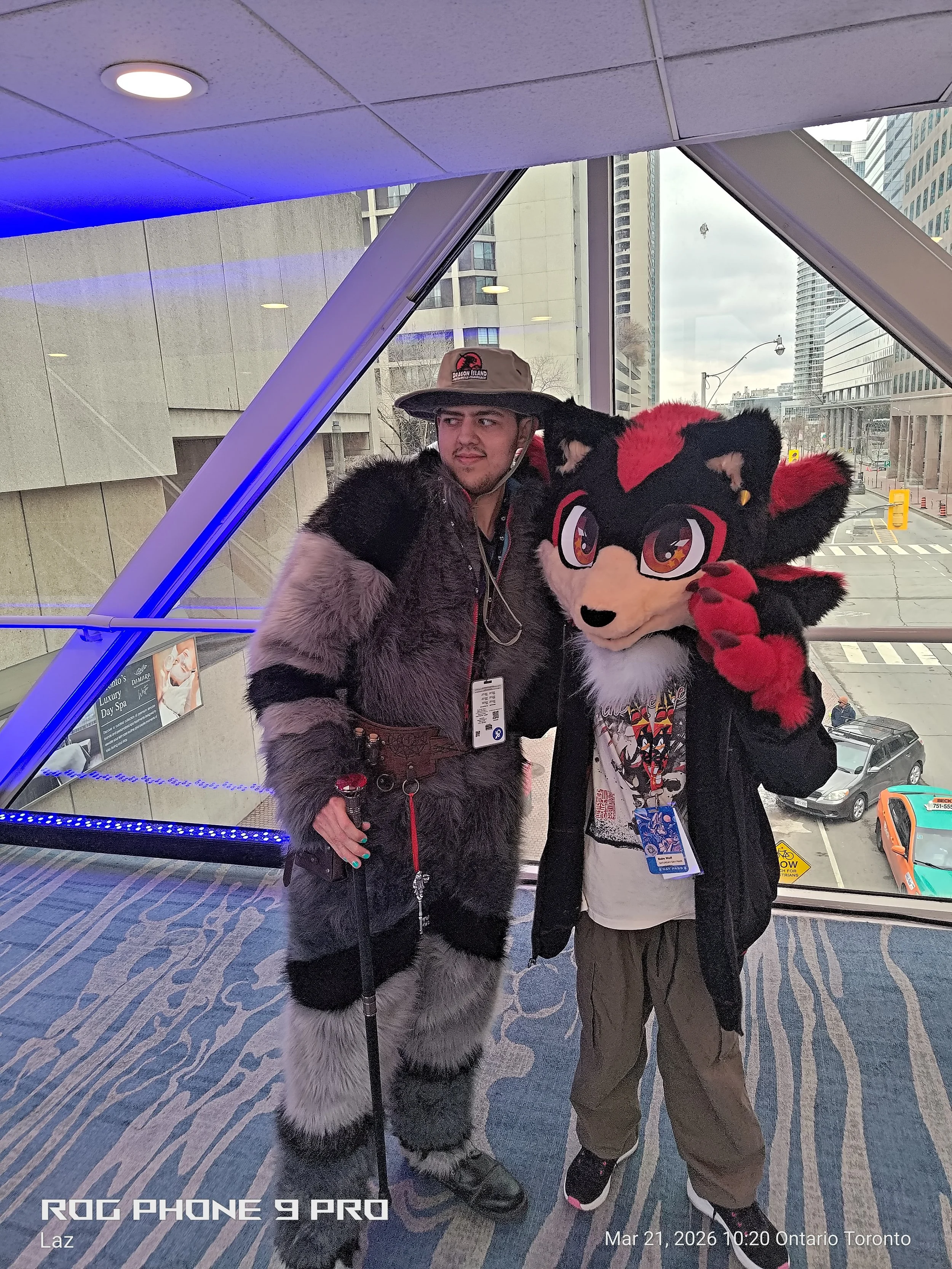 A person dressed in a full furry costume of a wolf-like character with black, red, and white fur, standing next to a man in casual clothing with a furry coat and a hat, on a blue patterned carpet in a modern building with large windows overlooking a 