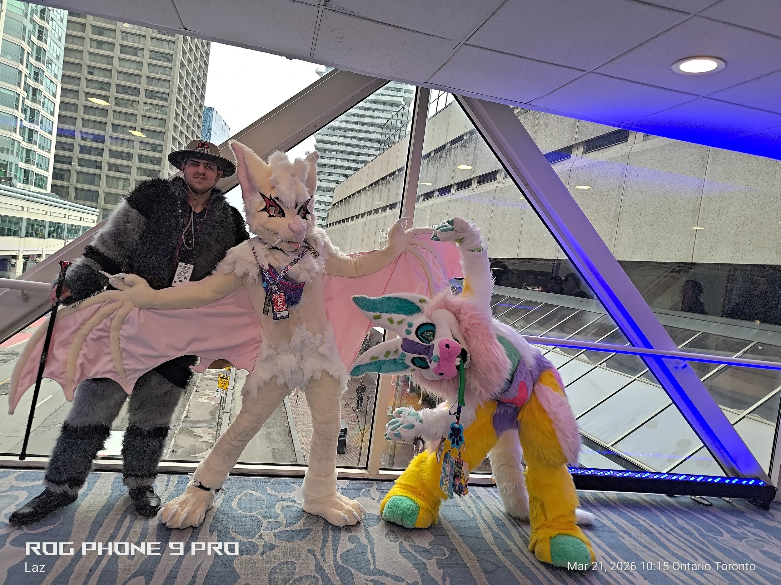 A man dressed in a black and gray furry costume and a person in a white fox costume with large eyes, a pink nose, and detailed facial features, standing inside a building with large glass windows. In the background, there are tall buildings and a cit