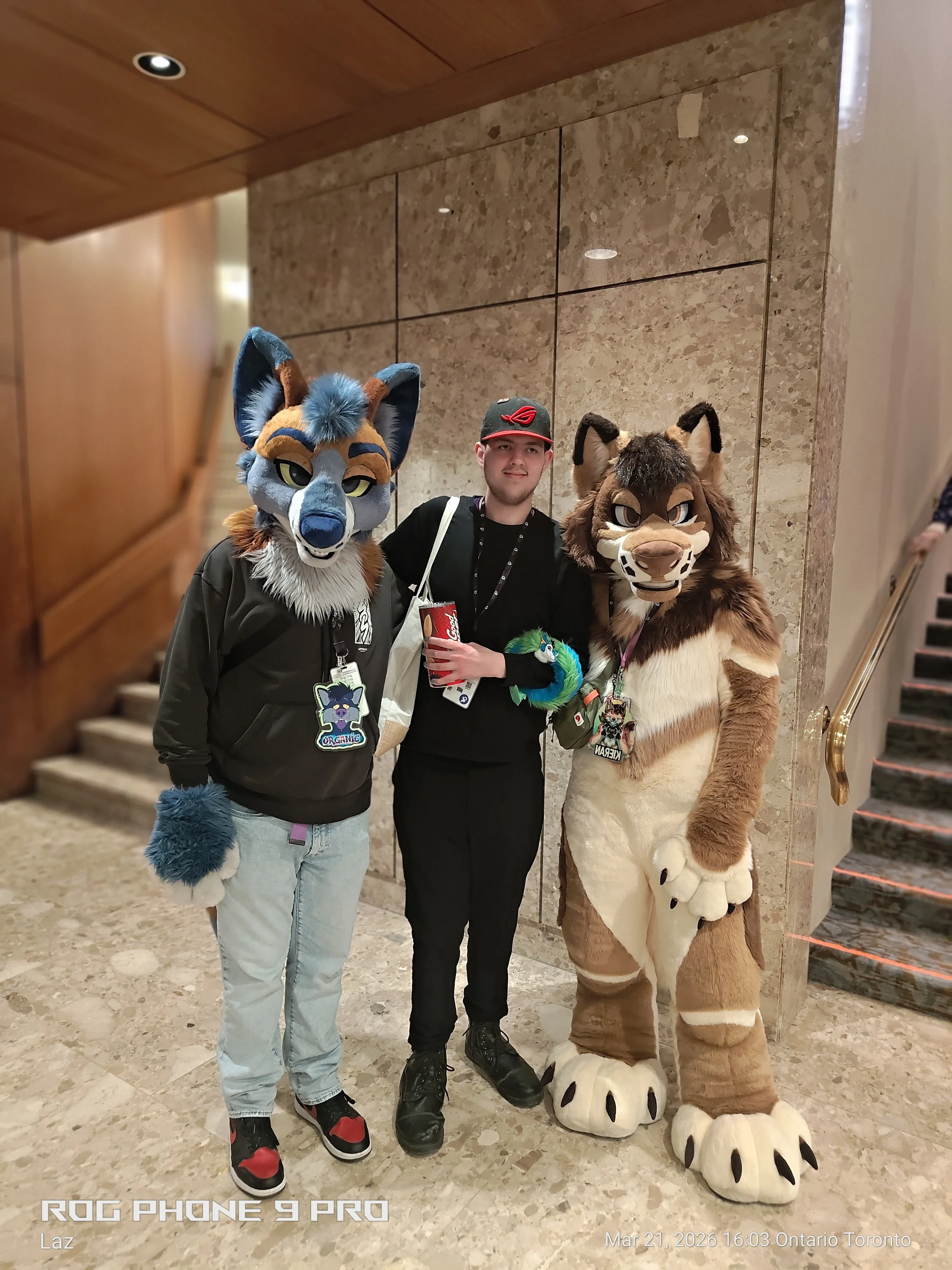 A man stands between two people in animal costumes, one in a blue and tan fox costume and the other in a brown and tan lion costume, inside a building with marble walls and stairs, holding a drink and smiling.