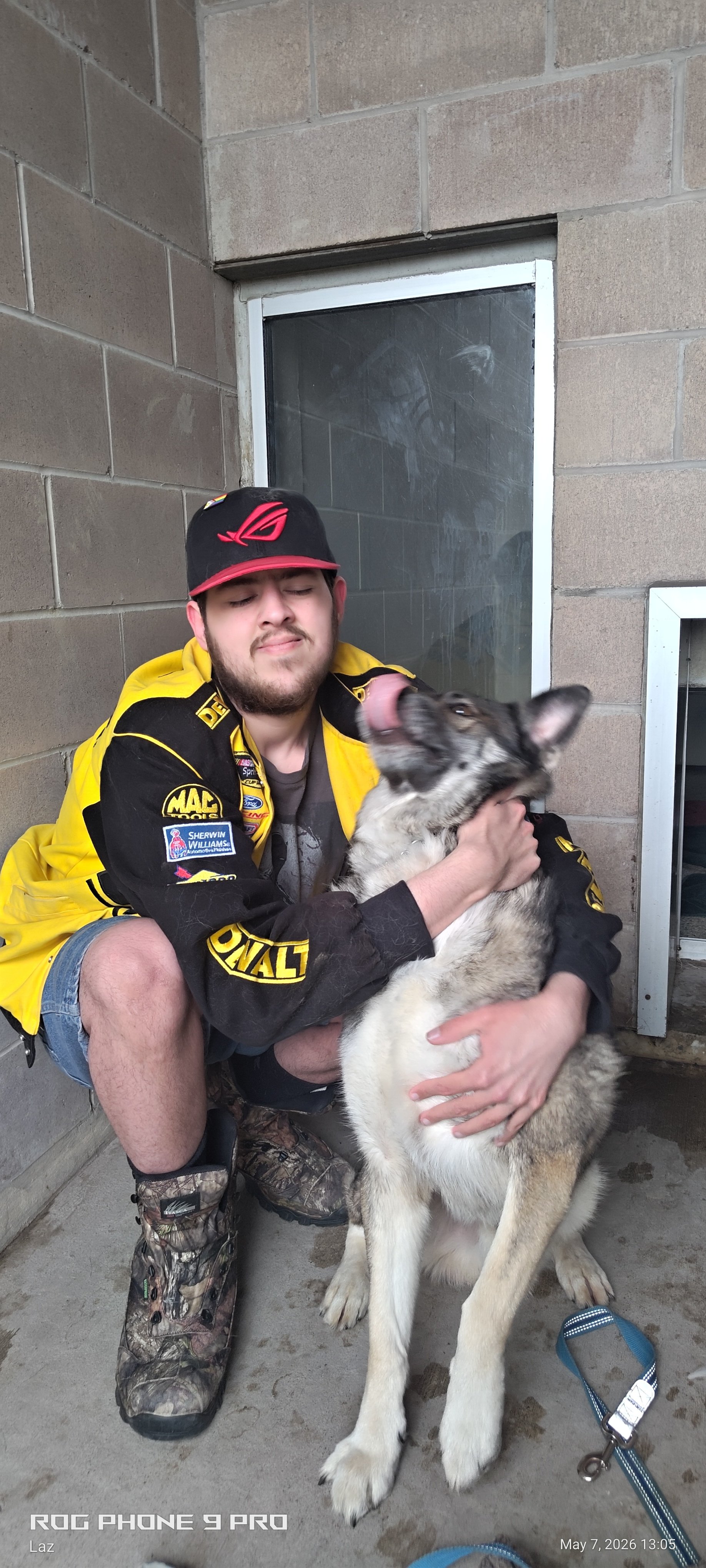 A young man in a racing jacket and camo boots squatting and hugging a large Siberian husky dog.