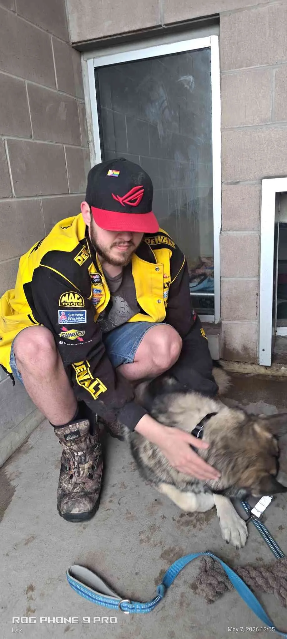 A man wearing a racing jacket, camouflage boots, and a cap, sitting on the ground and petting a dog, with a concrete wall, a small window, and a locker in the background.