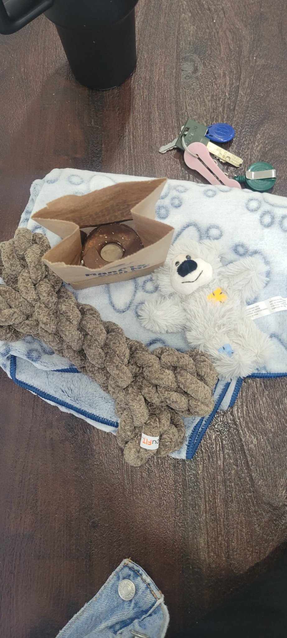 A brown paper bag with a donut inside, a plush white teddy bear, a thick rope, a gray and white towel, a set of keys, a blue and green key fob, a plastic cup, and part of a person's blue denim jeans and a black shoe.