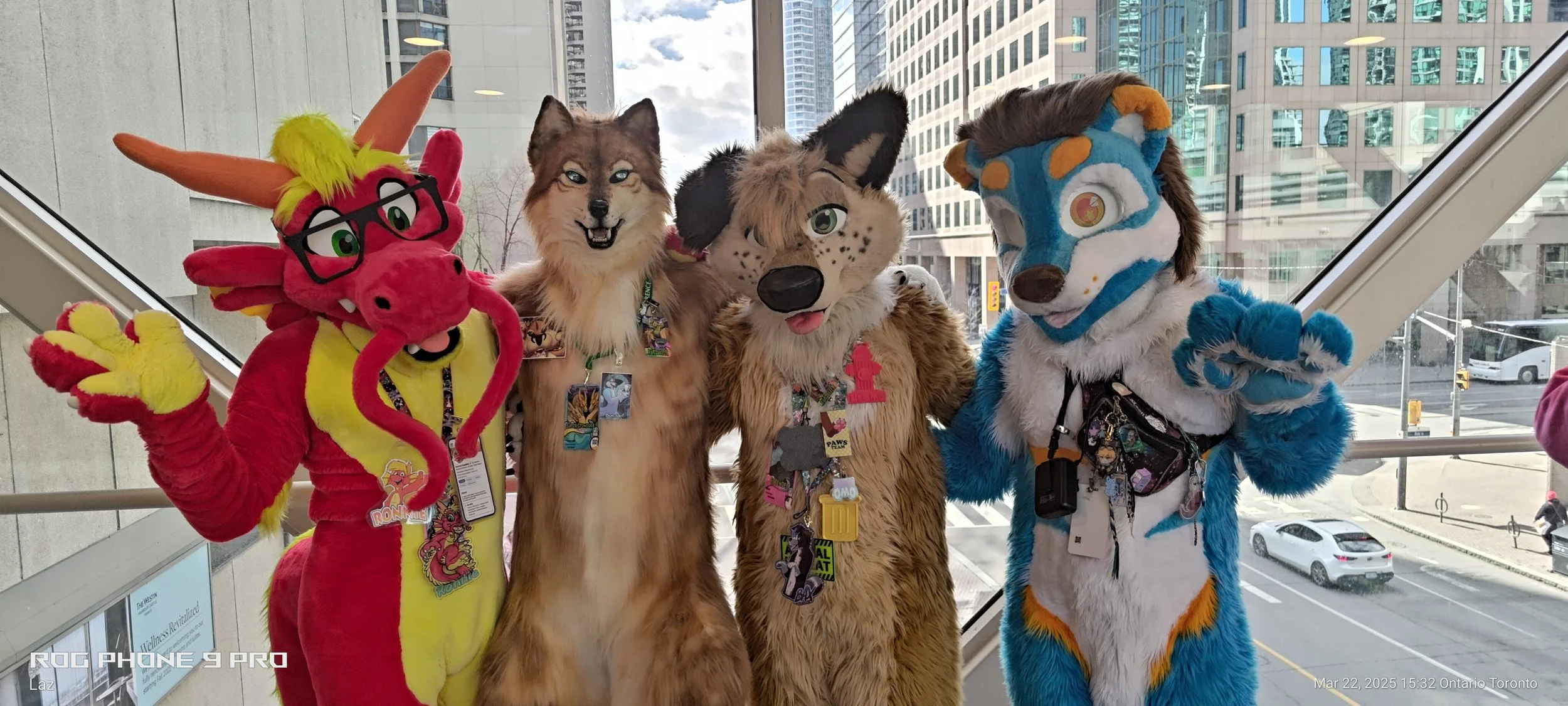 Four people in colorful animal costumes, including a red dragon with glasses, a wolf, a cheetah, and a blue and white dog, standing together inside a building with large windows.